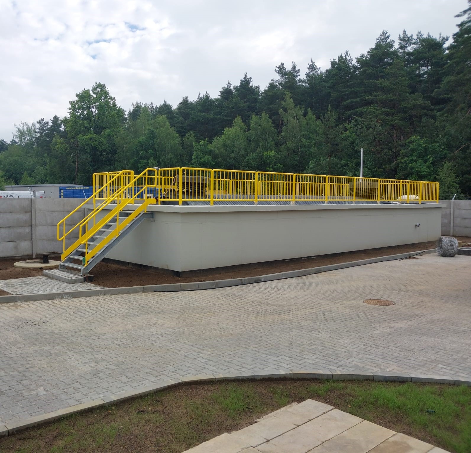 Zewnętrzny zbiornik retencyjny z żółtymi metalowymi schodami i balustradami, otoczony kostką brukową i zielenią, na tle lasu.