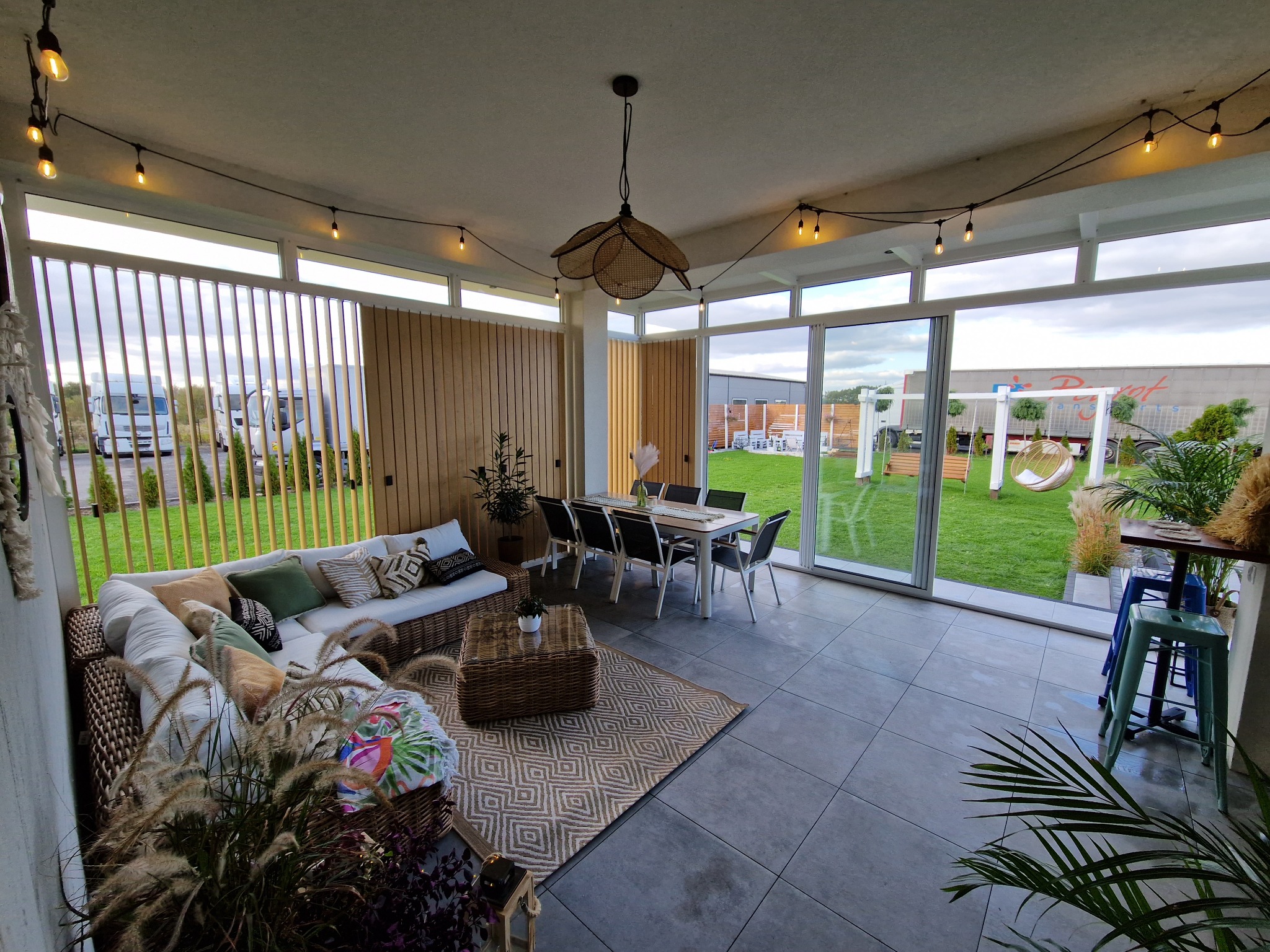 Przestronny, zadaszony taras w Rzeszowie z rattanowym wypoczynkiem, stołem jadalnym i widokiem na zielony ogród z huśtawką.