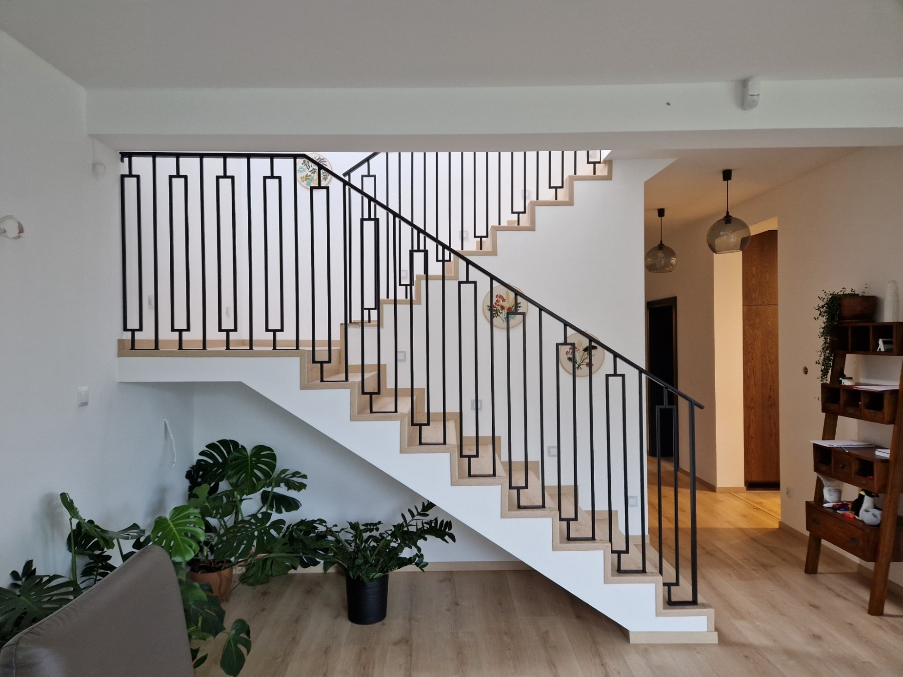 Nowoczesne schody z drewnianymi stopniami i czarną, metalową balustradą o geometrycznym wzorze, doświetlone małymi lampkami w ścianie, z dekoracjami roślinnymi w tle.