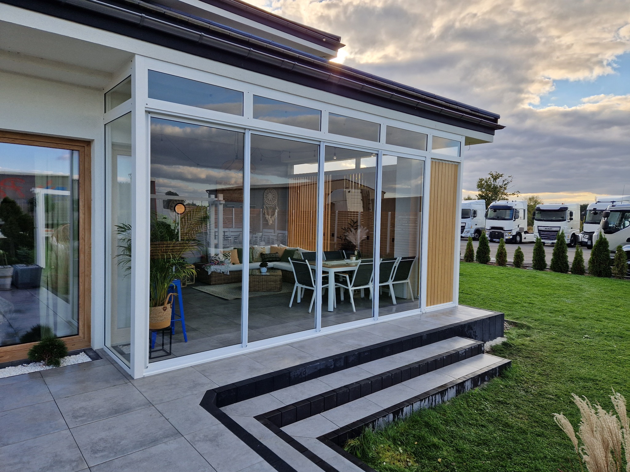 Nowoczesna przeszklona weranda z przesuwnymi drzwiami, widok na wnętrze z meblami ogrodowymi, schody wejściowe z ciemnymi obrzeżami, na zewnątrz trawnik i zaparkowane samochody dostawcze w tle.