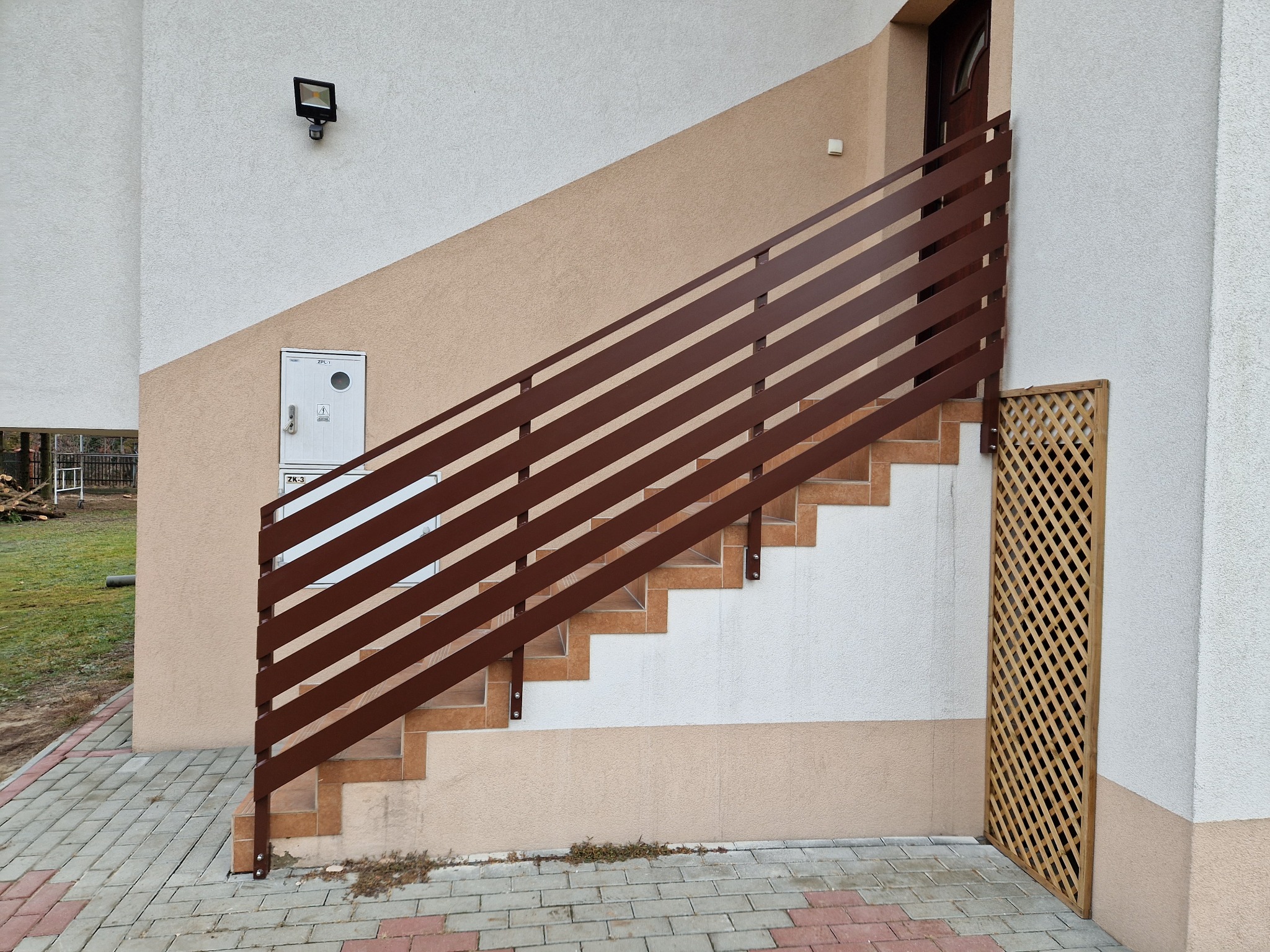 Zewnętrzne schody z brązową, poziomą balustradą z metalowych profili, prowadzące do drzwi wejściowych budynku z beżowo-białą elewacją.