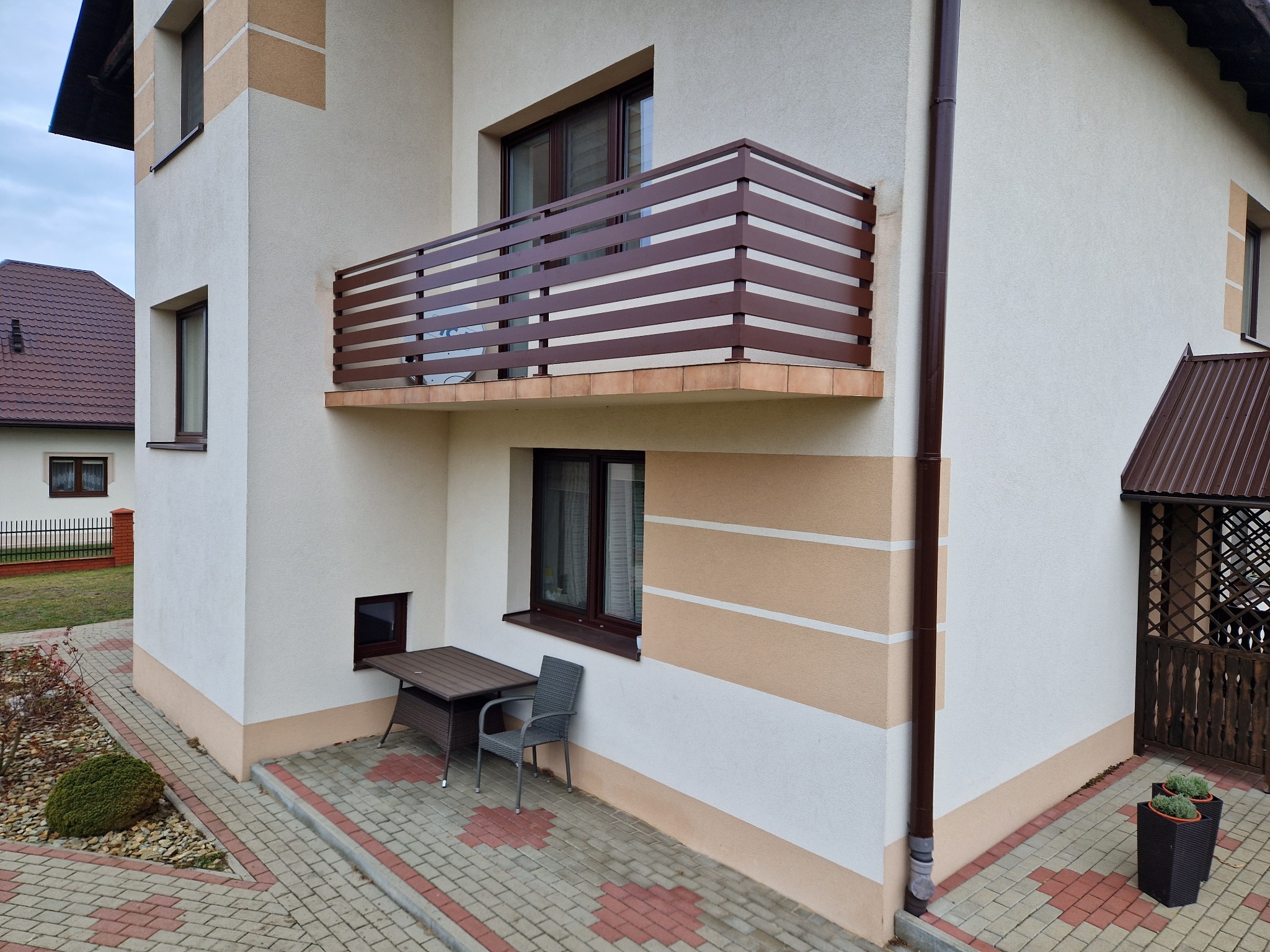 Brązowa, pozioma balustrada balkonowa z prostokątnych profili na elewacji domu w jasnych barwach, widok z dołu na balkon i fragment elewacji, na dole stolik i krzesło.
