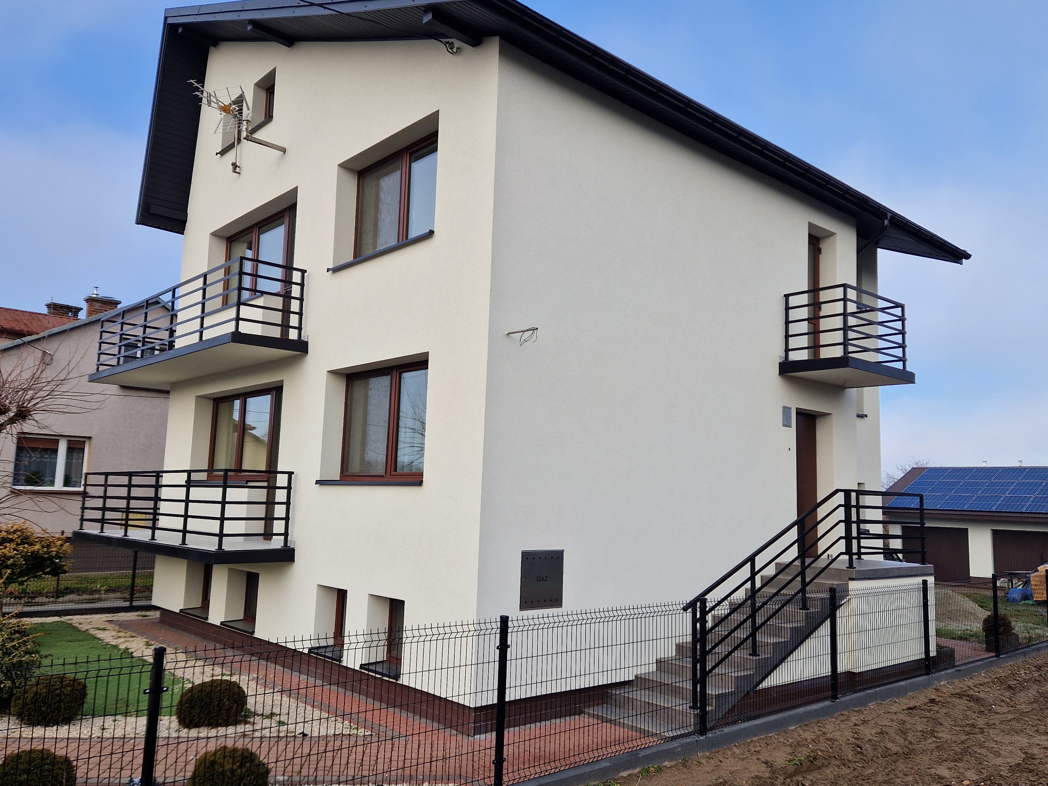 Nowoczesny dom dwupiętrowy z białymi ścianami, czarnym dachem i balkonami z czarnymi metalowymi balustradami, widoczne schody wejściowe z poręczą i ogrodzenie posesji z siatki.