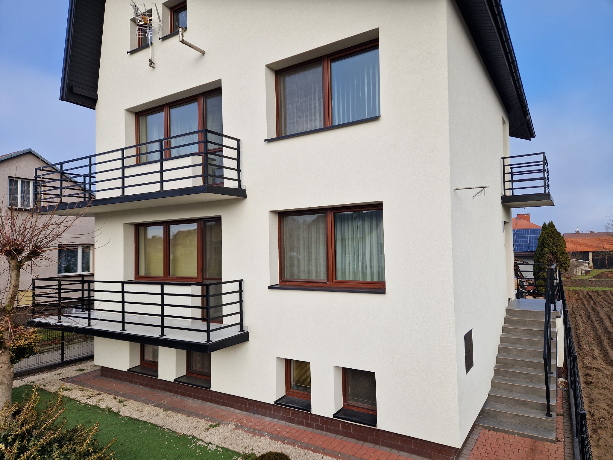 Nowoczesny, trzypiętrowy dom w Rzeszowie z balkonami z czarnymi, metalowymi barierkami. Widoczne schody zewnętrzne z poręczą prowadzące na taras.