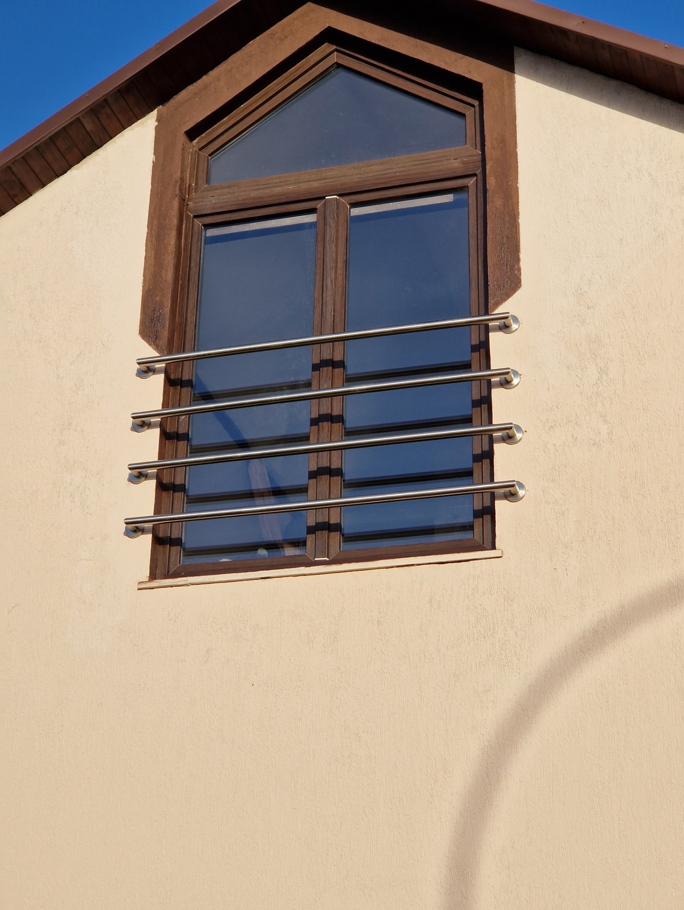 Okno na poddaszu z brązową ramą i zabezpieczeniem w postaci poziomych, metalowych prętów mocowanych do ramy za pomocą okrągłych uchwytów.