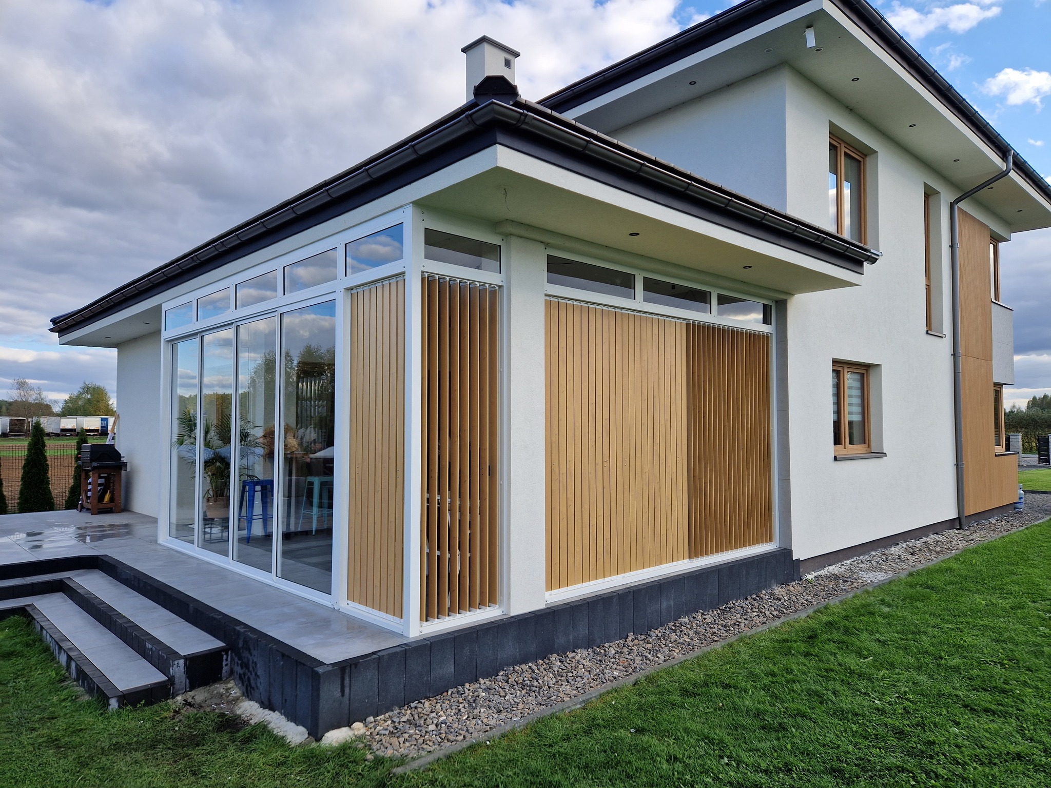 Nowoczesna dobudówka do domu z dużymi przesuwnymi oknami i drewnianymi żaluzjami pionowymi, z tarasem wyłożonym płytkami i schodami, otoczona trawnikiem i kamieniami.