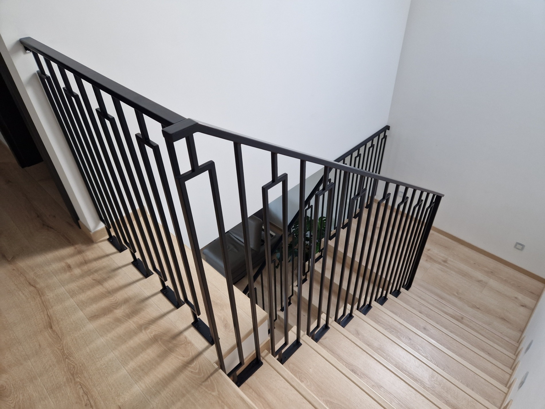 Nowoczesna, czarna, metalowa balustrada schodowa z geometrycznym wzorem, widok z góry na drewniane schody w jasnym wnętrzu.