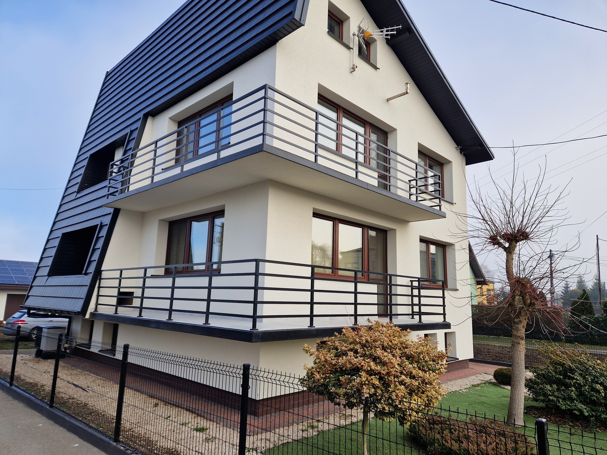 Nowoczesny dom z balkonami zabezpieczonymi czarnymi, prostymi balustradami, widok z dołu na elewację z kontrastującym, ciemnym dachem i jasnymi ścianami.