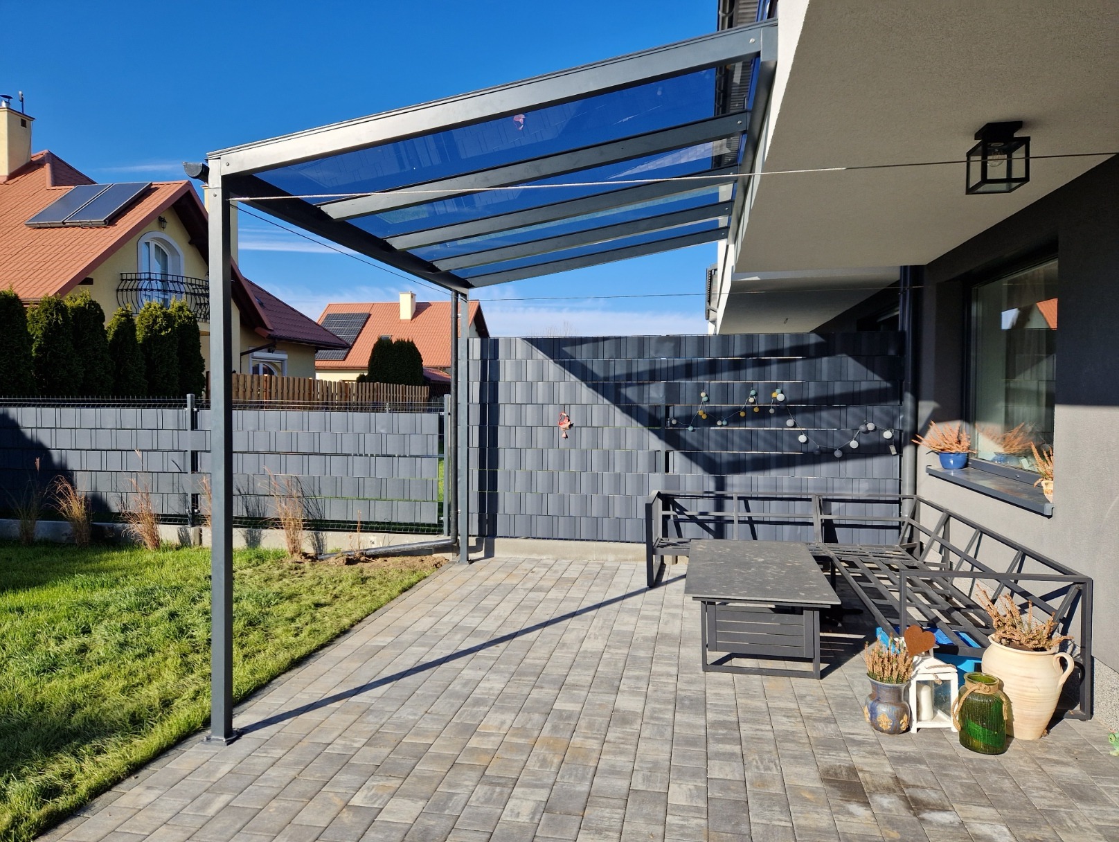 Metalowa pergola z przezroczystym zadaszeniem na tarasie z szarej kostki, obok metalowa ławka i stół, w tle szary płot panelowy i dom z czerwonym dachem.
