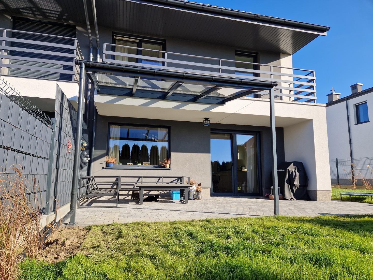 Nowoczesny dom z szarą elewacją, balkonem z metalową balustradą i przeszkloną pergolą nad tarasem wyłożonym kostką brukową, widok z trawnika w słoneczny dzień.