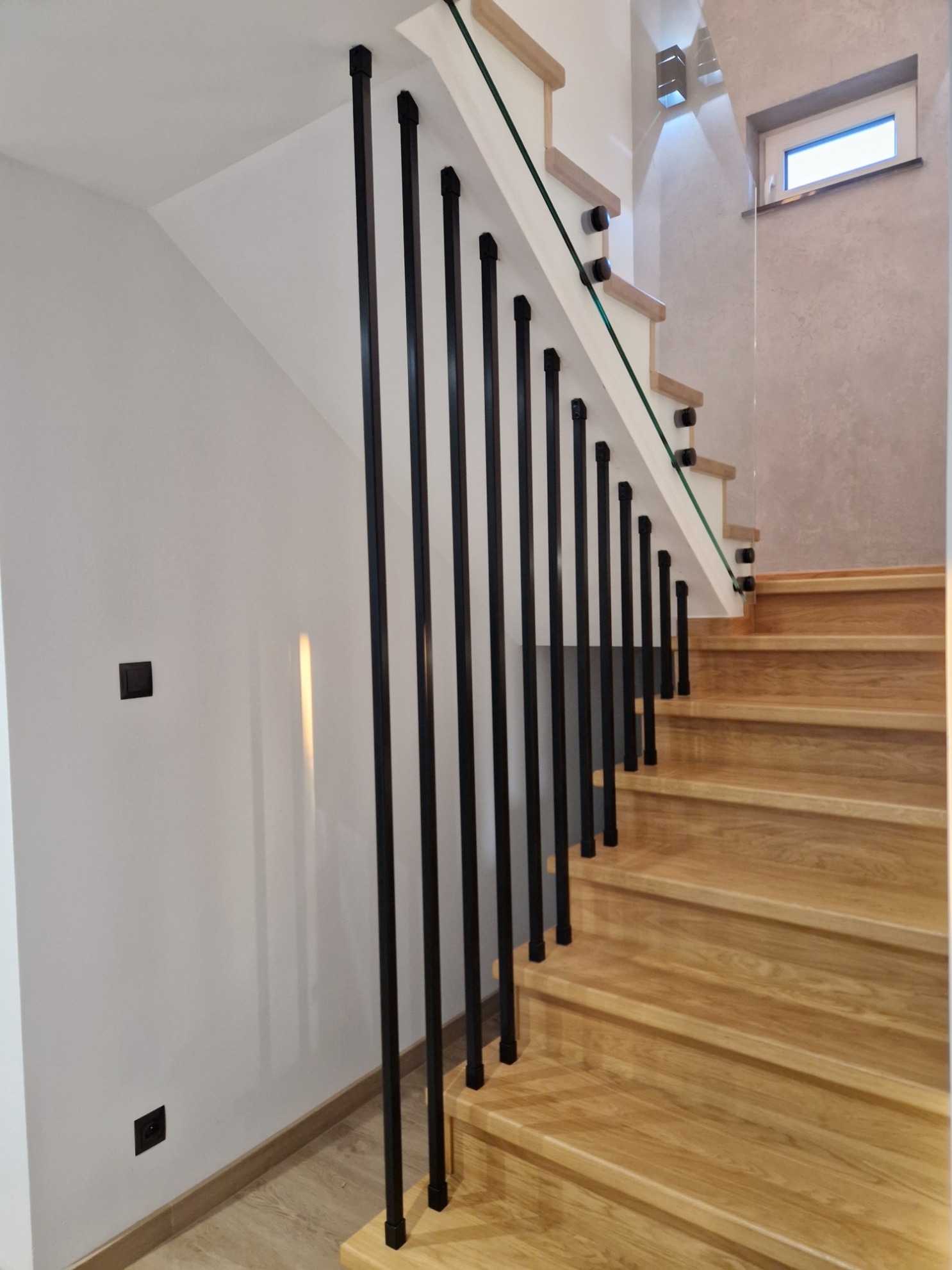 Jasne, drewniane schody z czarnymi, pionowymi słupkami balustrady i szklanym panelem, widok z dołu, oświetlenie punktowe na ścianie.
