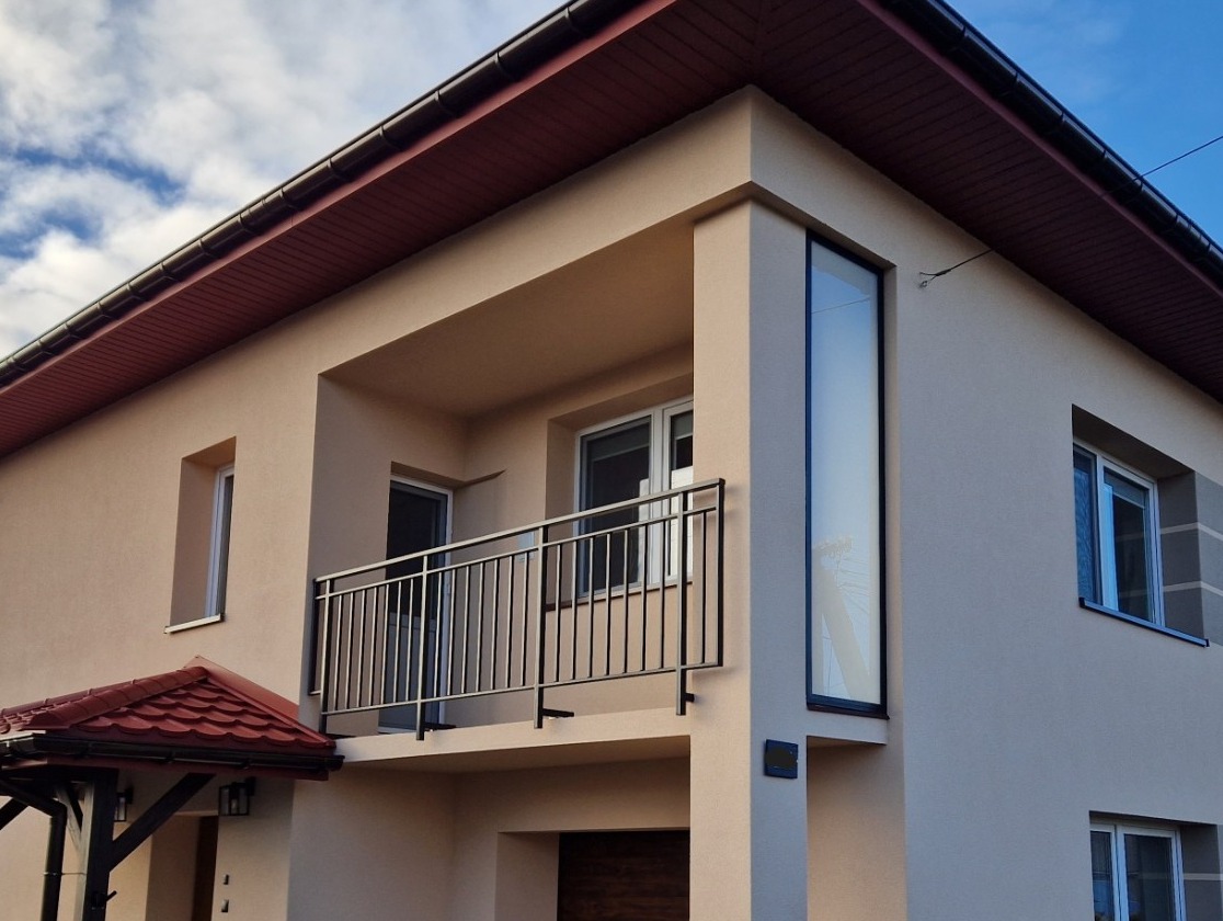 Nowoczesny dom z beżową elewacją, balkonem z czarną metalową balustradą i wysokim oknem narożnym.