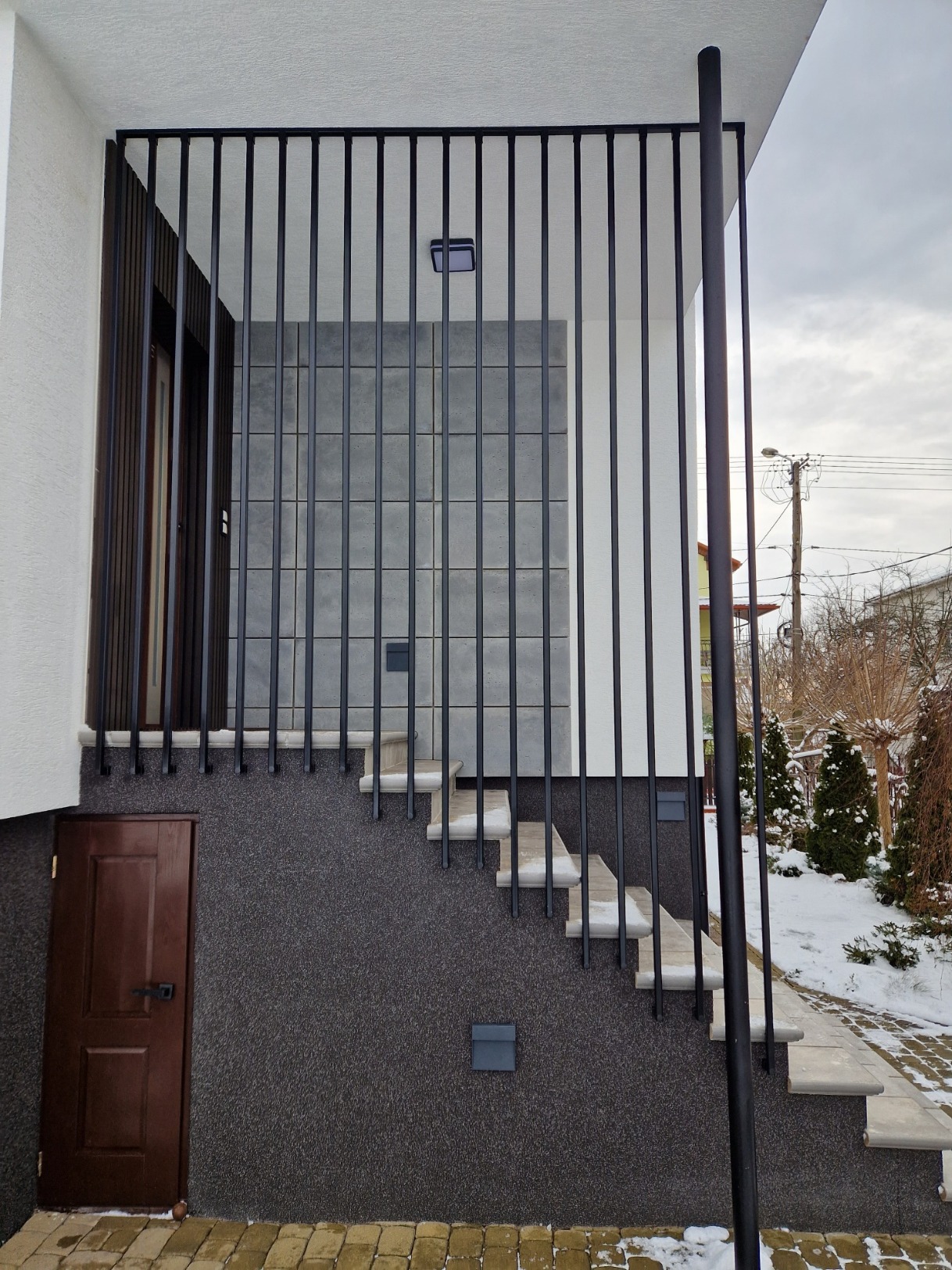 Nowoczesna, czarna balustrada metalowa z pionowych prętów zabezpieczająca schody wejściowe do domu, z widocznymi elementami wykończenia elewacji i fragmentem zimowego krajobrazu w tle.