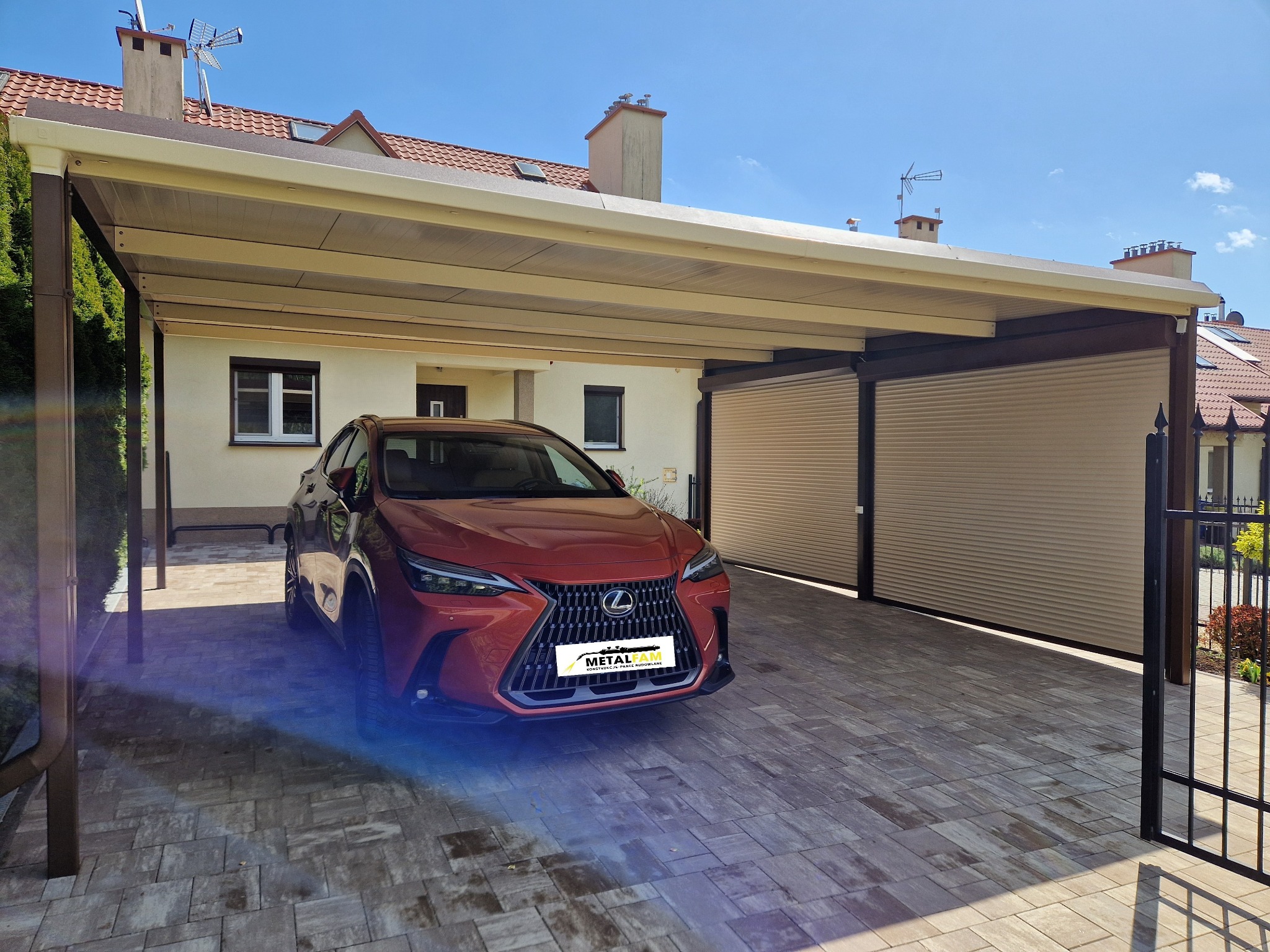 Nowoczesna, brązowo-beżowa wiata garażowa z roletami, chroniąca czerwony samochód Lexus na kostce brukowej, z widocznym fragmentem ogrodzenia i domu w tle.