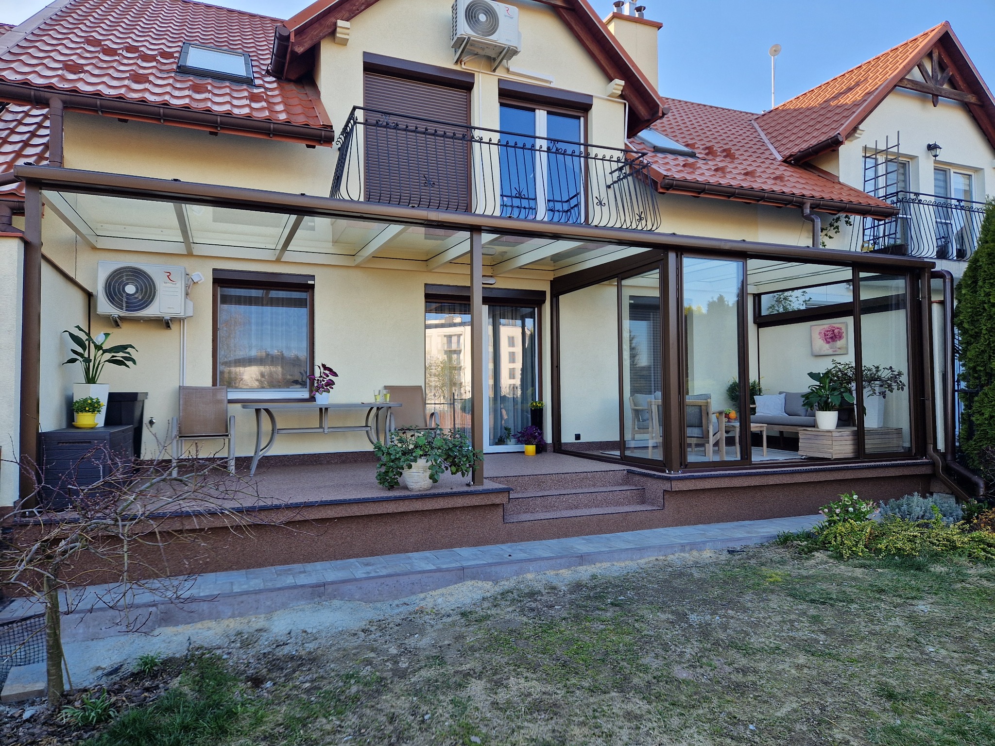 Zadaszony taras z przeszklonymi ścianami w brązowej ramie, widok z zewnątrz na jasny salonik z sofą i roślinami doniczkowymi, w tle dwupiętrowy dom z balkonami i czerwoną dachówką.