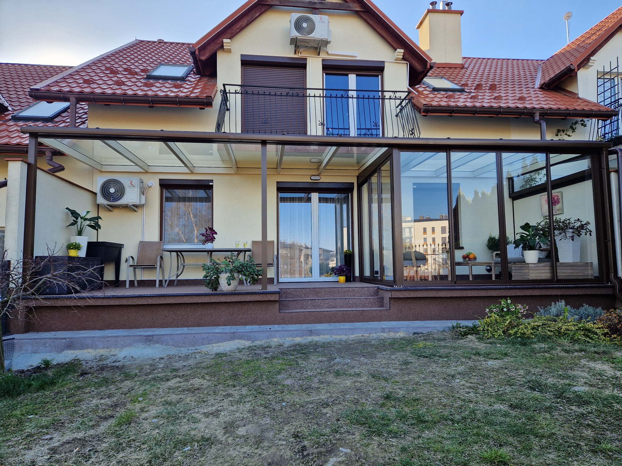 Zadaszony taras z przeszklonymi ścianami i szklanym dachem, przylegający do domu z balkonem i brązową balustradą. Widoczny stół z krzesłami i roślinami w doniczkach na tarasie. Zewnętrzny...