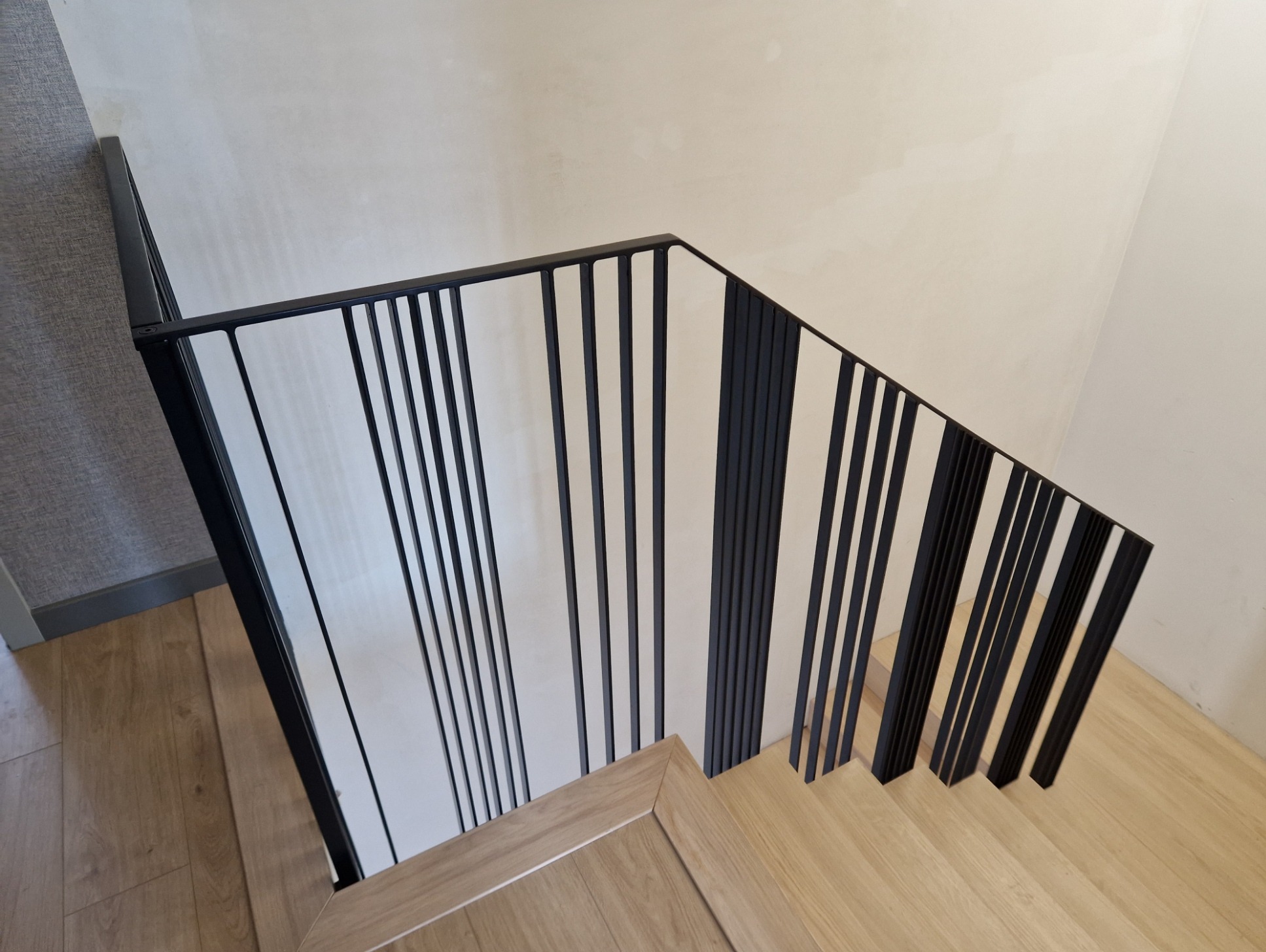 Nowoczesne schody z drewnianymi stopniami i czarną, minimalistyczną balustradą z pionowych prętów, widziane z góry pod kątem.