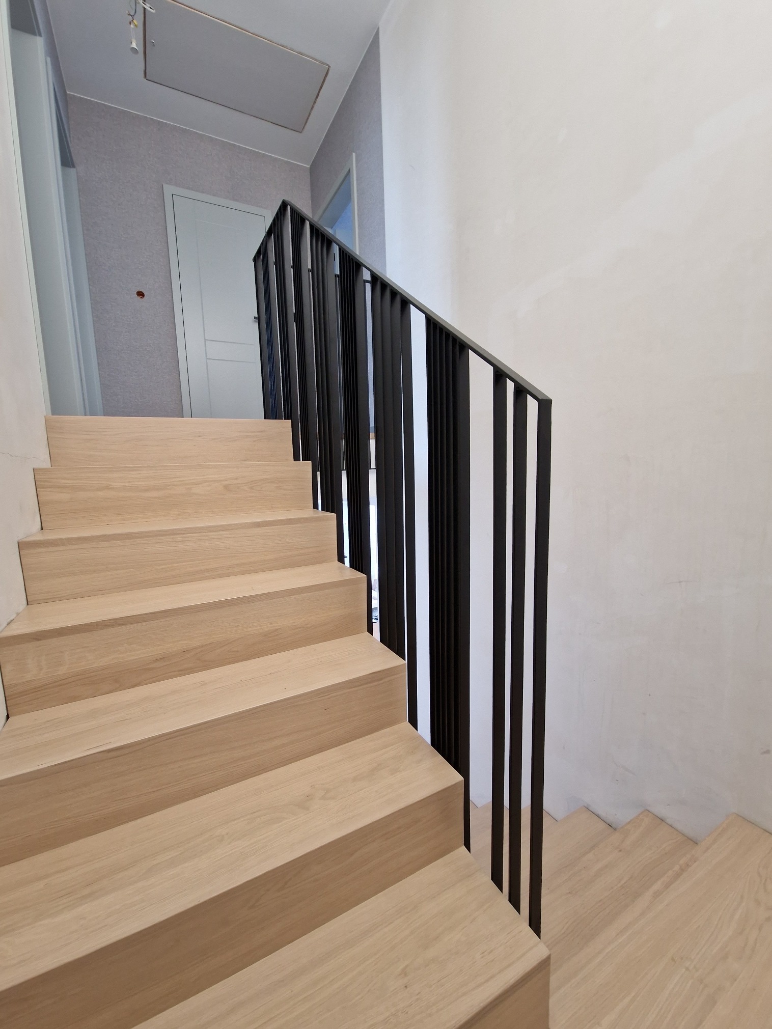 Jasne drewniane schody z czarną, minimalistyczną balustradą w nowoczesnym wnętrzu. Widoczne proste, pionowe elementy balustrady i fragment ściany.