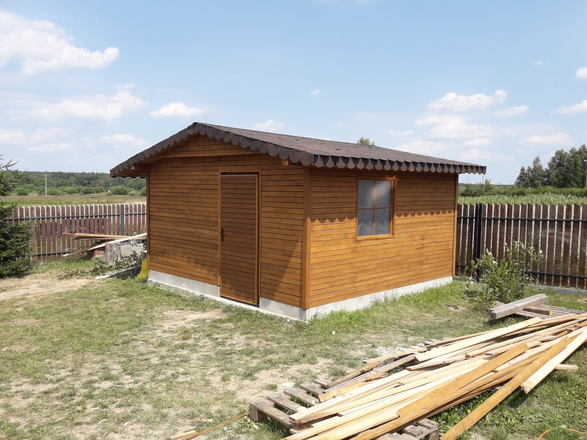 Nowo wybudowany, drewniany domek narzędziowy z brązowym dachem, widoczny na trawniku w słoneczny dzień, z ułożonymi obok deskami i paletą.