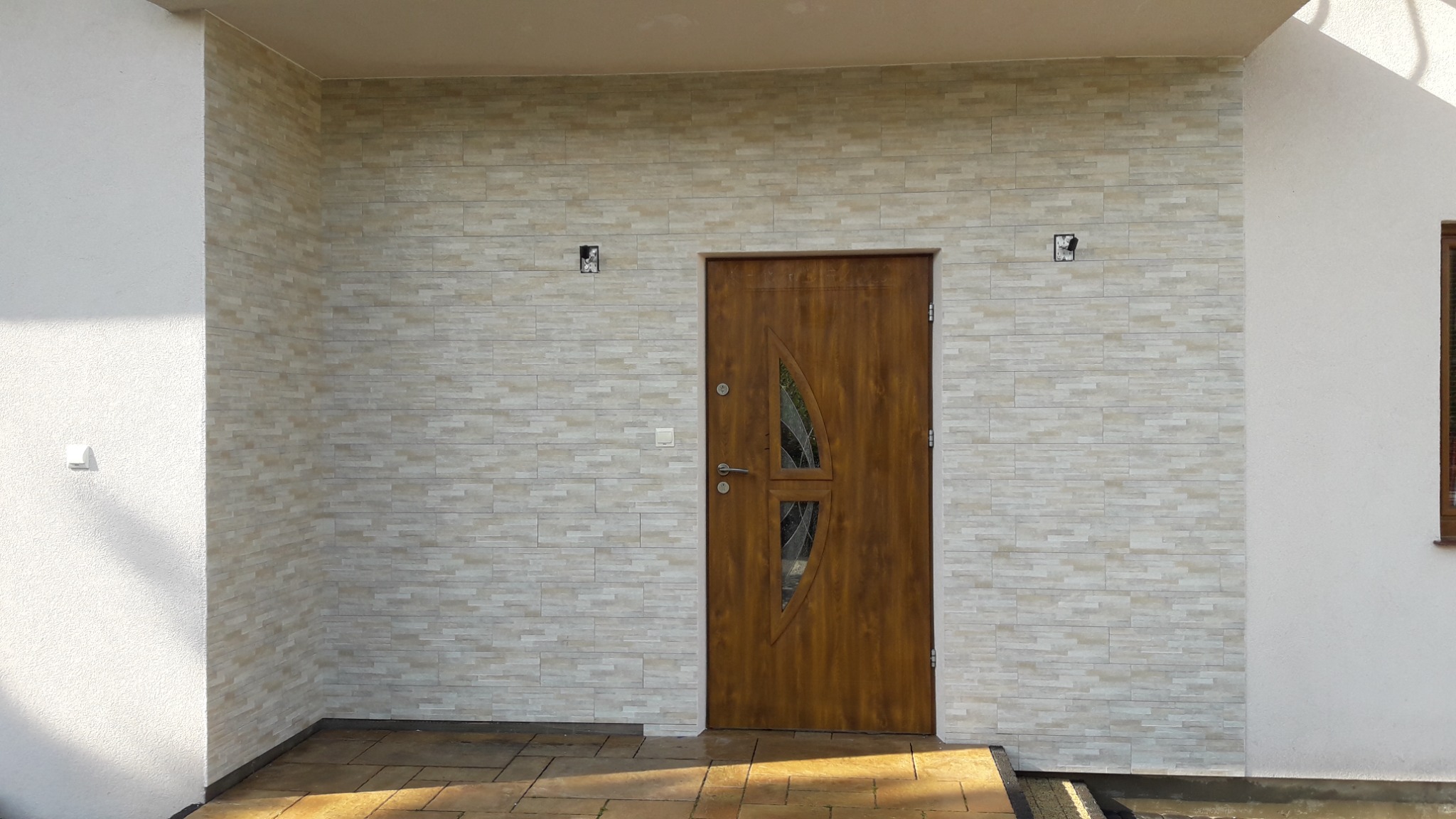 Elewacja domu wykończona jasnymi płytkami imitującymi kamień, z brązowymi drzwiami wejściowymi z przeszkleniami o nietypowym kształcie.