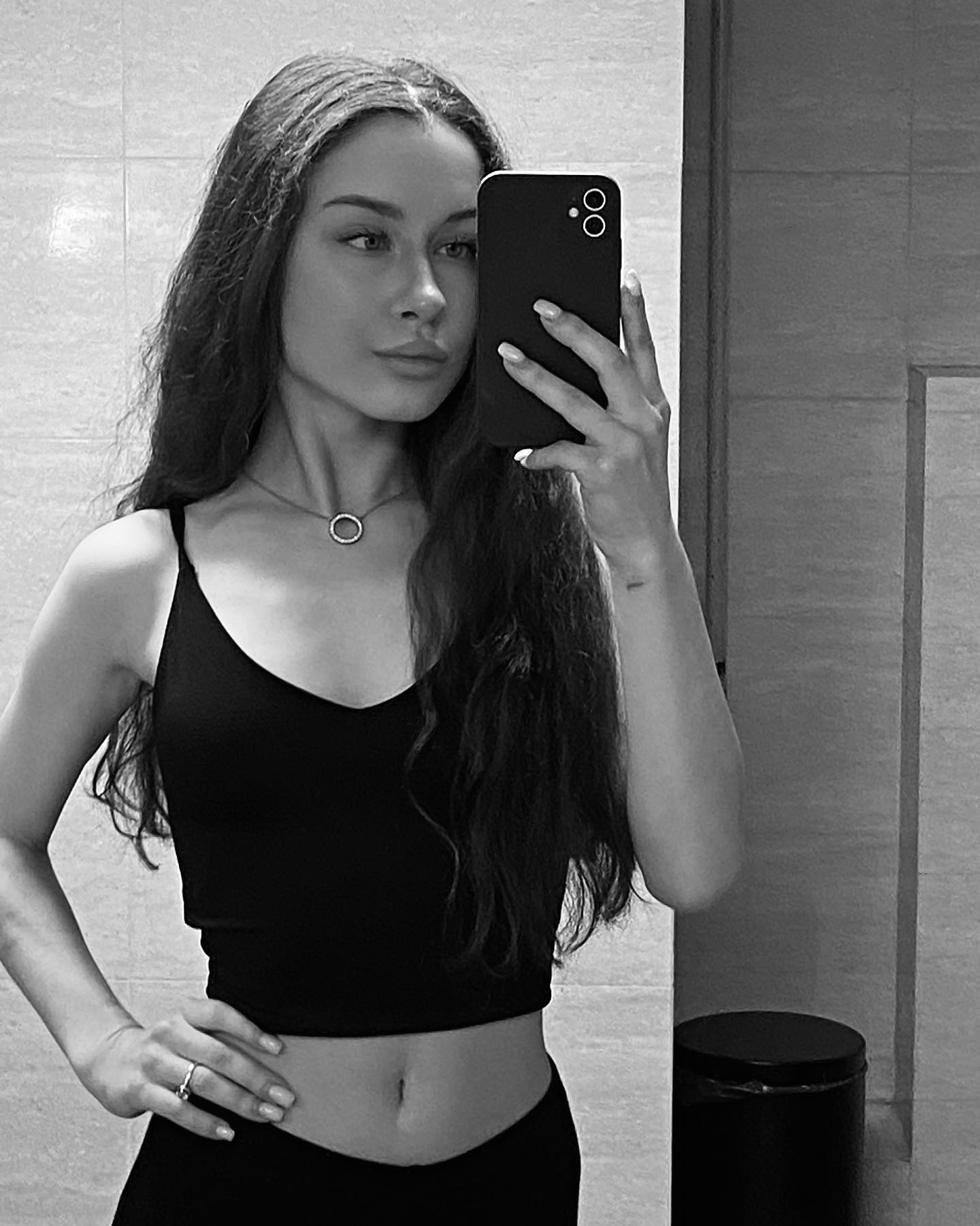Czarno-białe selfie młodej kobiety w czarnym topie i spodniach, z długimi falowanymi włosami, srebrnym naszyjnikiem i pierścionkiem, pozującej w łazience z telefonem w dłoni.