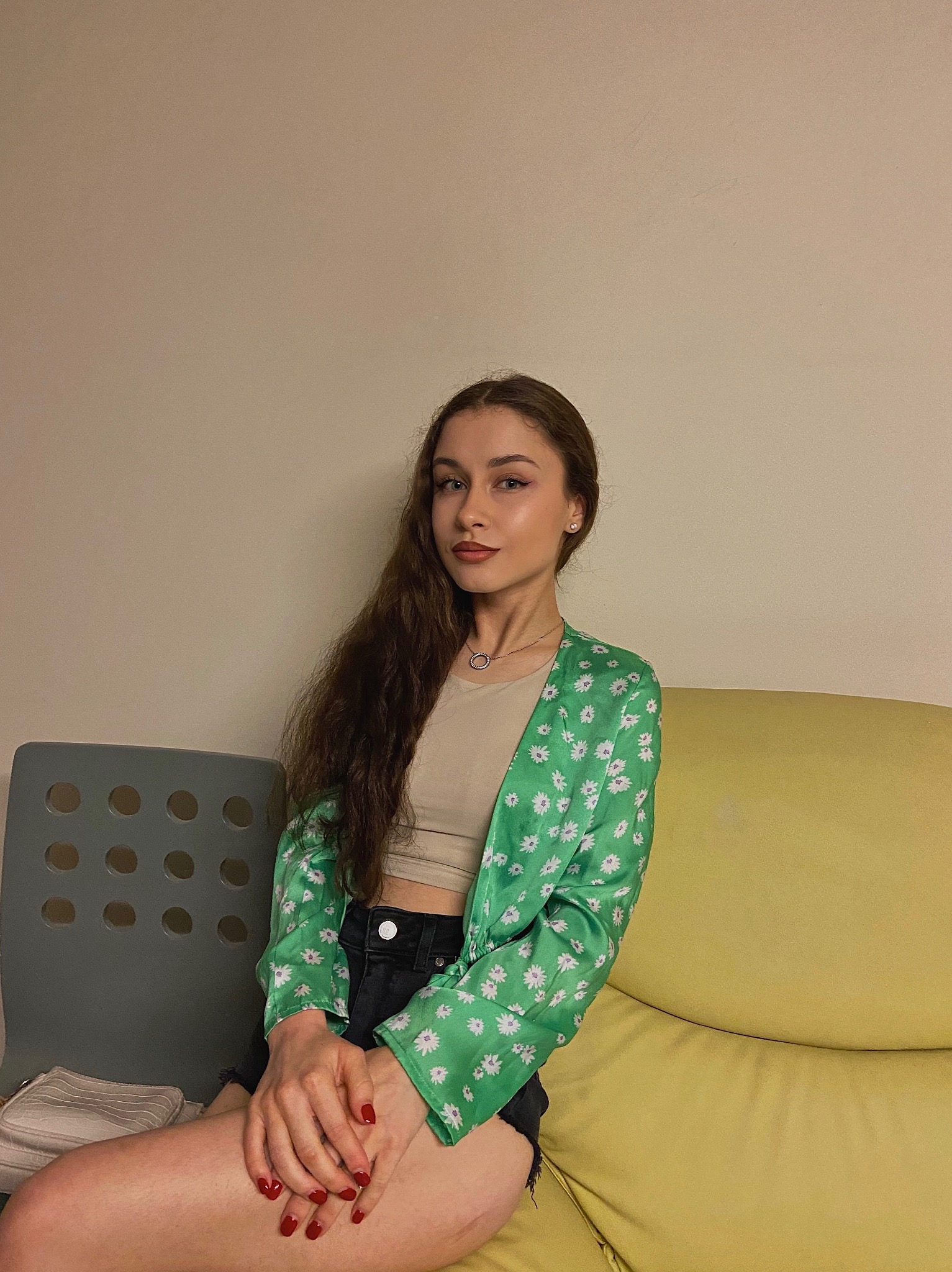 Portret młodej kobiety siedzącej na żółtej sofie, ubrana w zieloną bluzkę w białe kwiaty, czarne szorty i beżowy top, z długimi brązowymi włosami i czerwonymi paznokciami.