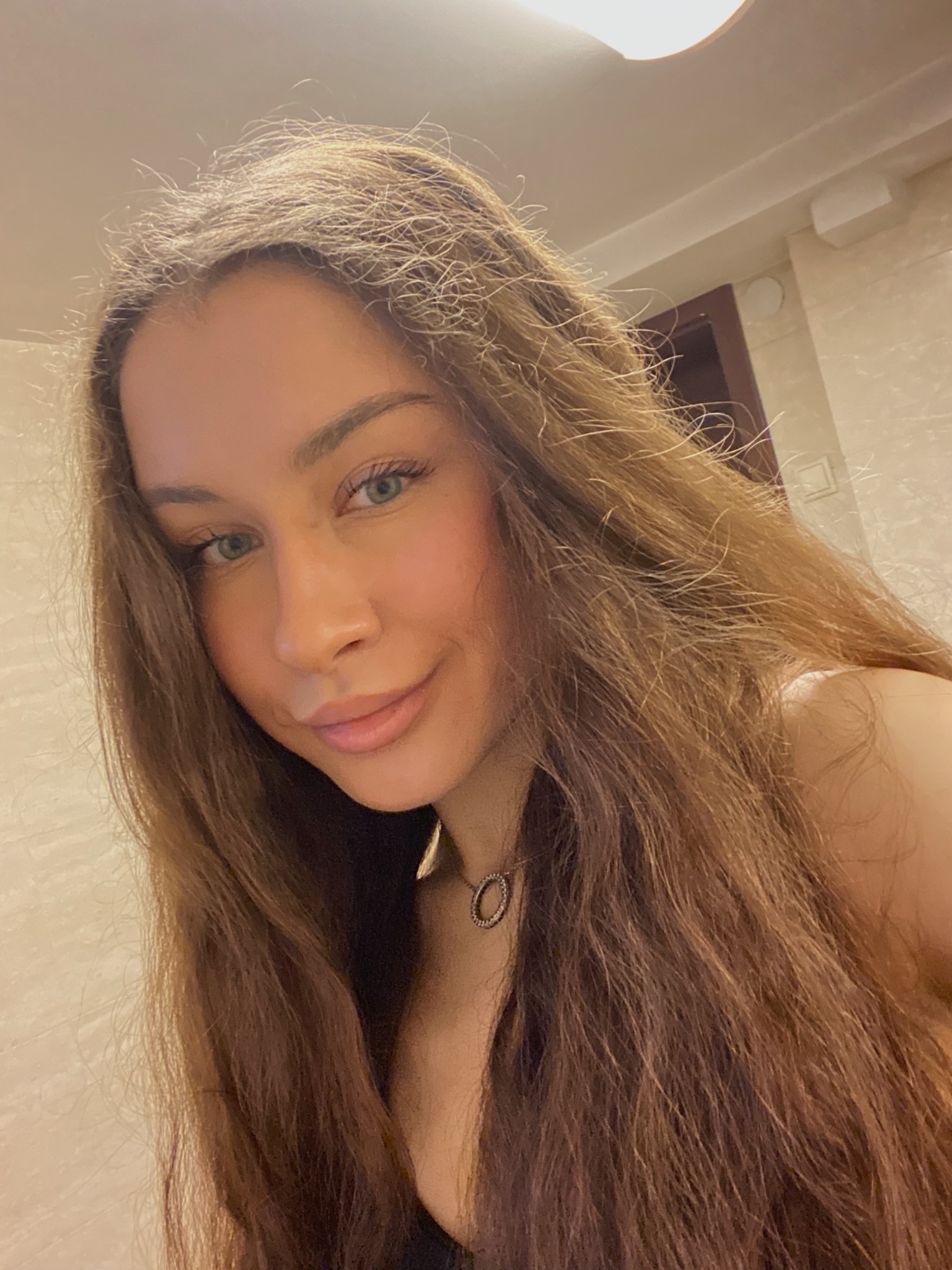 Portret młodej kobiety z długimi brązowymi włosami i subtelnym naszyjnikiem, wykonany w Łodzi, z widocznym oświetleniem sufitowym i jasną ścianą w tle.