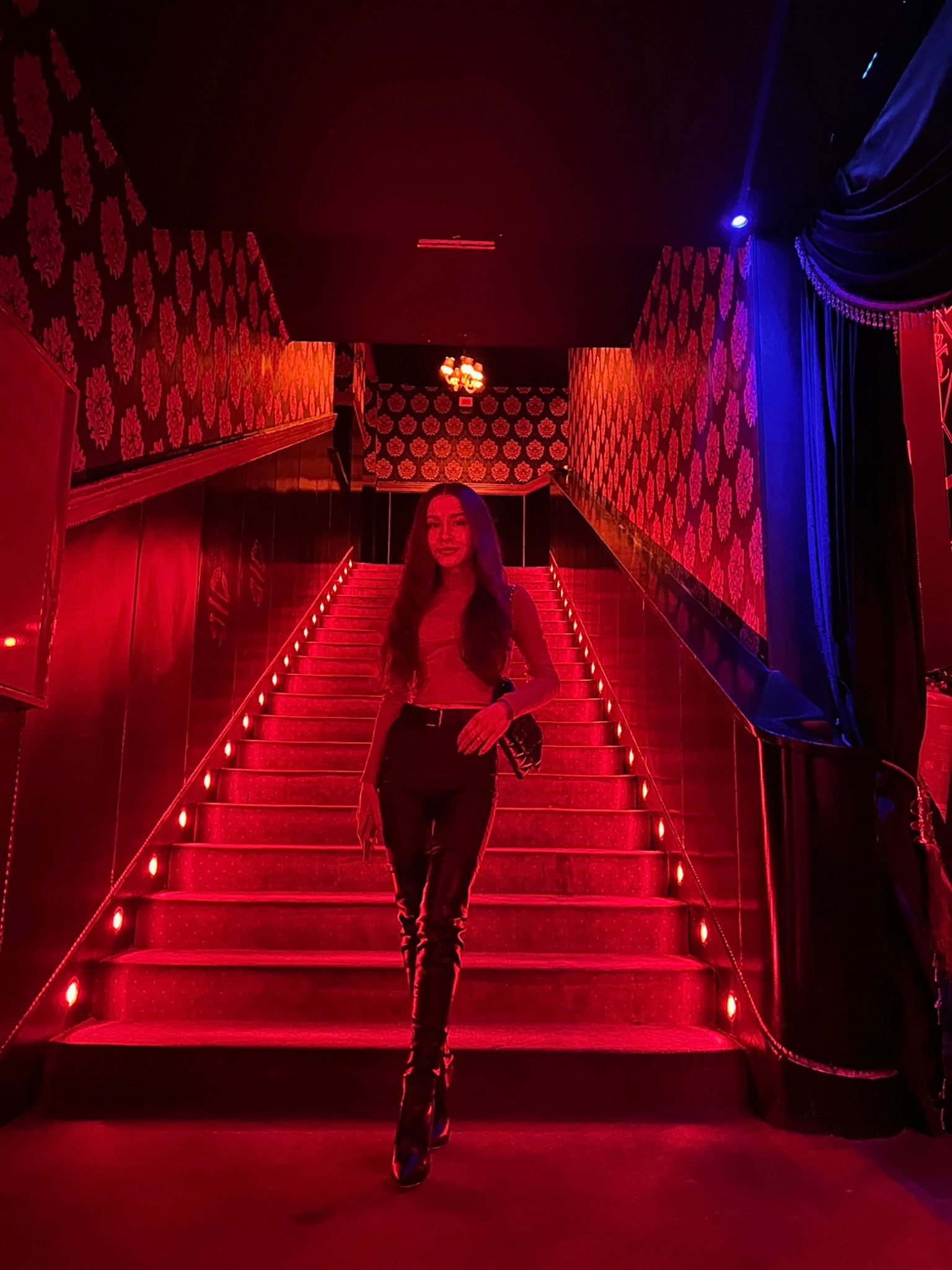 Kobieta w czarnych skórzanych spodniach i wysokich kozakach schodzi po czerwonych schodach z oświetleniem punktowym, tło stanowi wzorzysta tapeta w odcieniach czerwieni i czerni, z elementami...