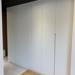 Meblospan Meble na wymiar - Duża, biała szafa wnękowa z minimalistycznymi, długimi uchwytami na tle ciemnego sufitu i drewnianej boazerii.