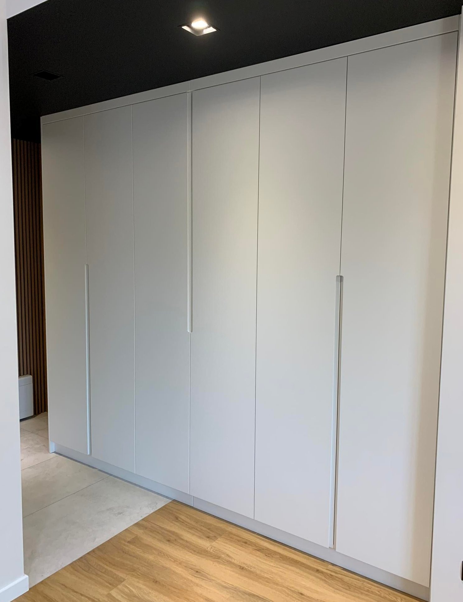 Duża, biała szafa wnękowa z minimalistycznymi, długimi uchwytami na tle ciemnego sufitu i drewnianej boazerii.