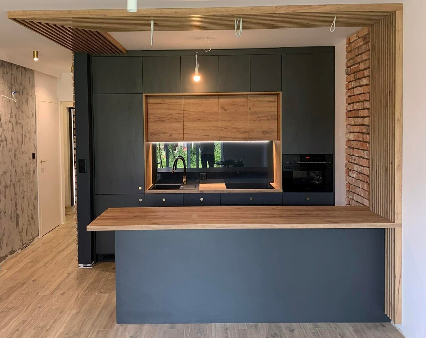 Nowoczesna kuchnia z ciemnoszarymi szafkami, drewnianymi akcentami i wyspą, oświetleniem LED pod szafkami i dekoracyjną ścianą z cegły oraz drewnianymi lamelami.