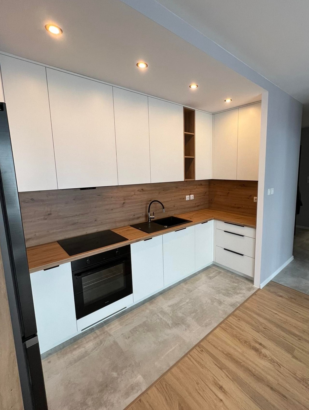 Nowoczesna kuchnia na wymiar z białymi frontami, drewnianym blatem i panelem ściennym, czarnym zlewem i płytą indukcyjną, oświetleniem punktowym.