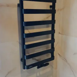 Czarny, drabinkowy grzejnik łazienkowy zamontowany na marmurowych płytkach ściennych, widoczne przyłącza.