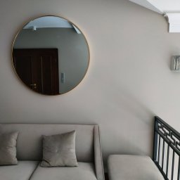 Sna Bud - Jasny salon z okrągłym lustrem w złotej ramie, odbijającym fragment ciemnych drzwi, oraz szarą sofą z poduszkami i metalową balustradą.
