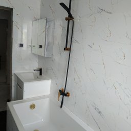 Sna Bud - Nowoczesna łazienka w trakcie wykańczania: biała wanna z złotymi odpływami, umywalka z szafką i czarną armaturą ze złotymi akcentami, ściany wyłożone białymi płytkami z marmurkowym wzorem.