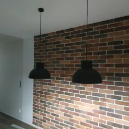 Ściana z dekoracyjnej cegły w odcieniach brązu i beżu, oświetlona dwoma czarnymi lampami wiszącymi w kształcie kloszy, widok na przedpokój z drzwiami wejściowymi.