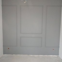 Sna Bud - Szara ściana z dekoracyjnymi panelami w kształcie prostokątów i kwadratów, widoczne gniazdka elektryczne, surowy beton na podłodze, stan surowy.