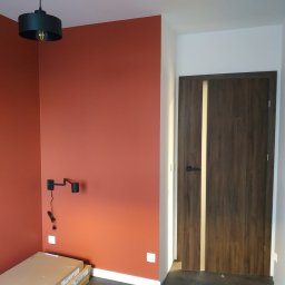 Sna Bud - Ściana pomalowana na rdzawy kolor z zamontowanym czarnym kinkietem, widoczna drewniana drzwi z jasnym paskiem, lampa sufitowa i kartony na podłodze.