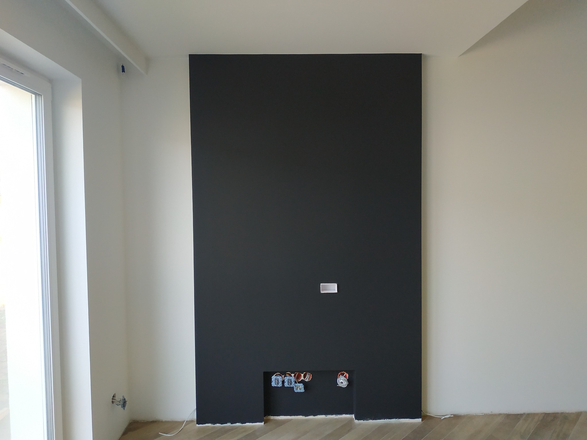 Ściana pomalowana na ciemny grafit z przygotowanymi otworami na gniazdka elektryczne i wnęką na przewody, stan surowy przed montażem osprzętu.