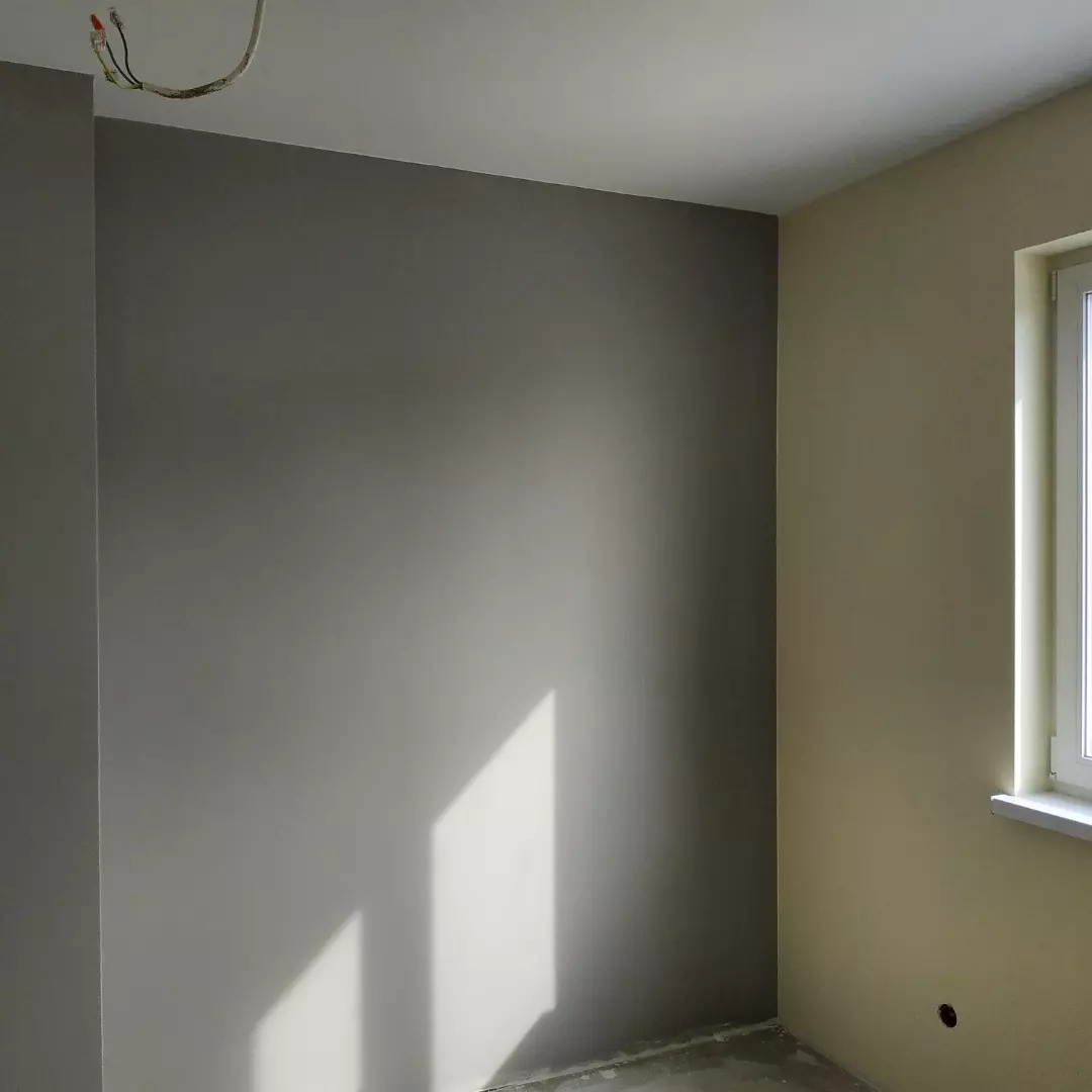 Pomieszczenie po malowaniu ścian na dwa kolory: szary i beżowy, z widocznym oknem i wystającymi kablami ze ściany.
