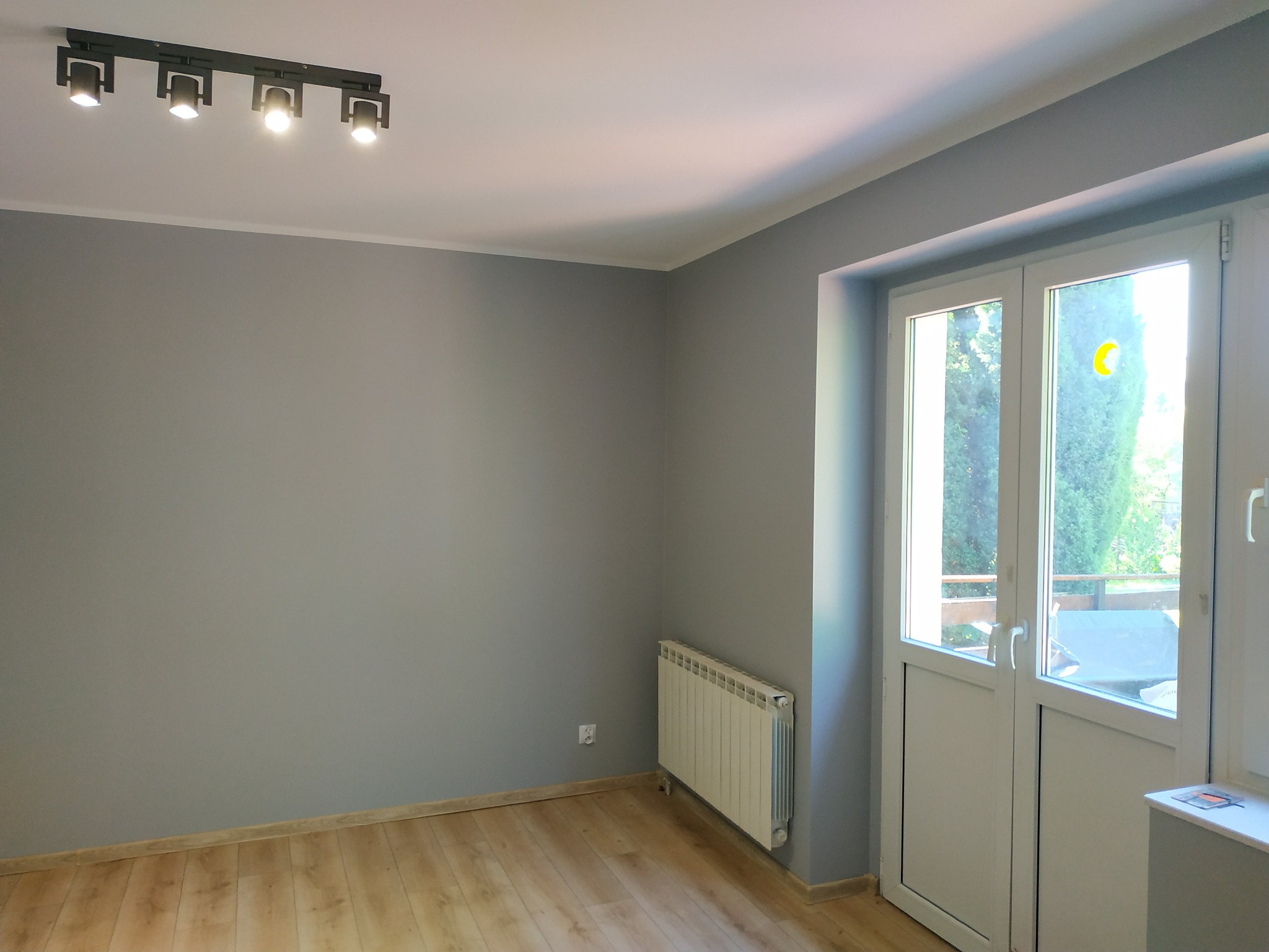 Wnętrze pokoju we Wrocławiu po remoncie: ściany pomalowane na szaro, drewniana podłoga, białe drzwi balkonowe z widokiem na zieleń, grzejnik i nowoczesne oświetlenie sufitowe.