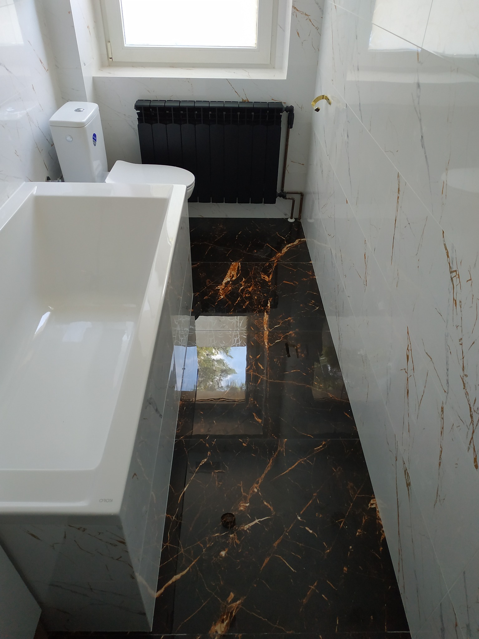 Nowoczesna łazienka z białą wanną KOLO, toaletą i czarnym grzejnikiem, wykończona marmurowymi płytkami w odcieniach bieli i brązu.