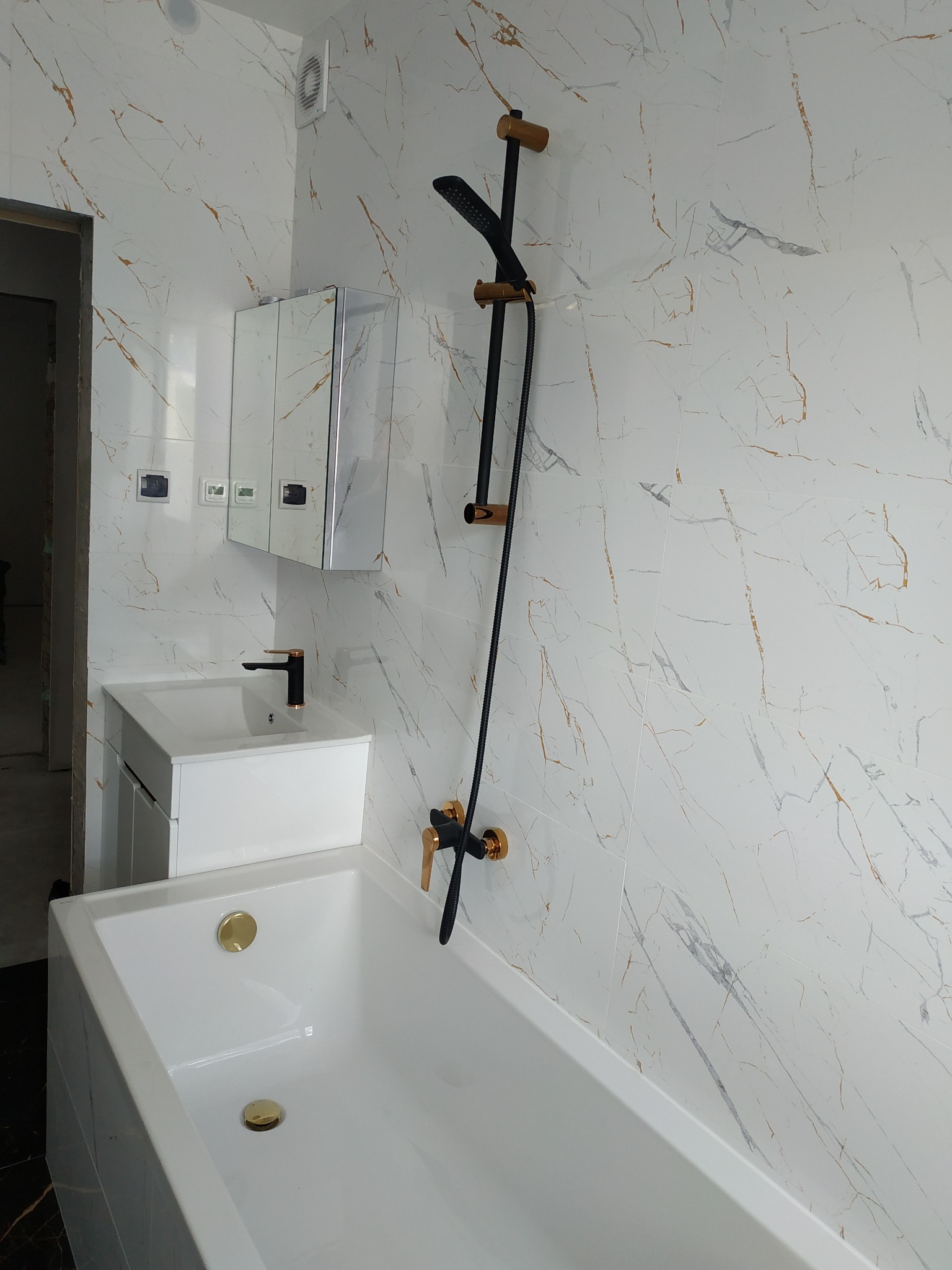 Nowoczesna łazienka w trakcie wykańczania: biała wanna z złotymi odpływami, umywalka z szafką i czarną armaturą ze złotymi akcentami, ściany wyłożone białymi płytkami z marmurkowym wzorem.