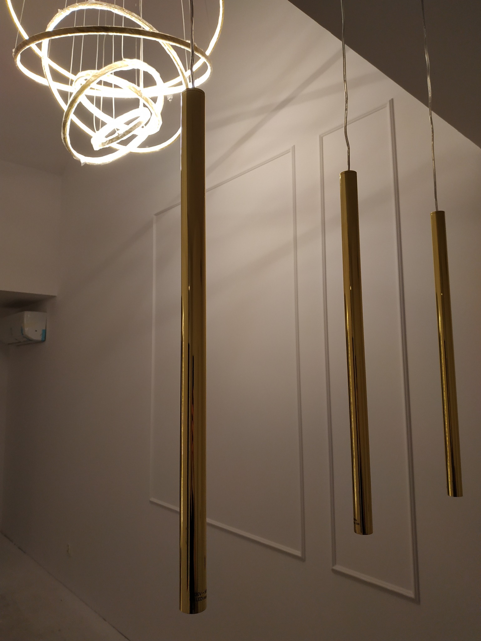 Nowoczesne oświetlenie wnętrza: złote, cylindryczne lampy wiszące na tle białej ściany z dekoracyjnymi panelami i okrągłym, wielopierścieniowym żyrandolem LED.
