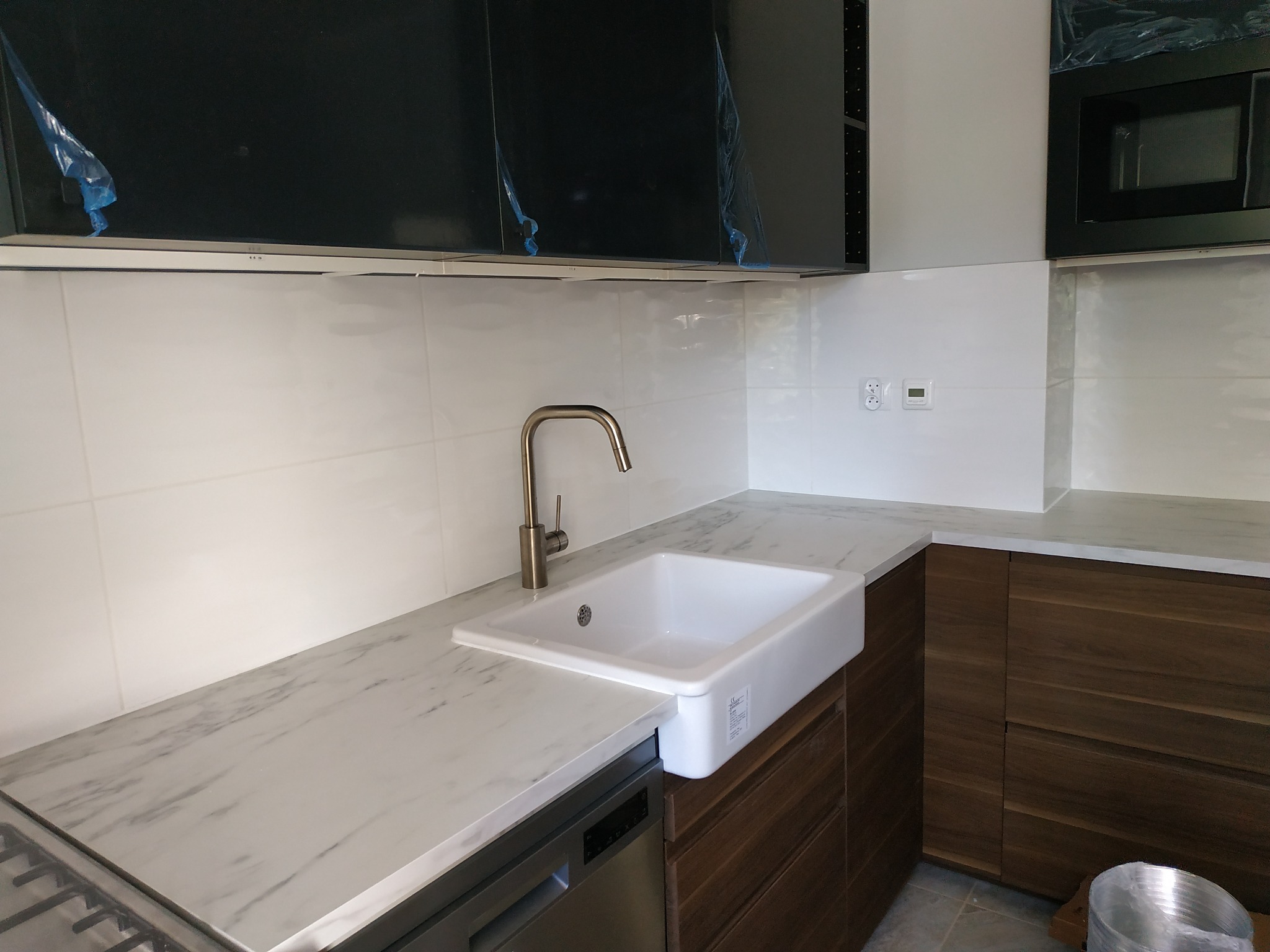 Nowoczesna kuchnia z białym, ceramicznym zlewem farmerskim, marmurowym blatem i drewnianymi szafkami; czarne szafki wiszące jeszcze w folii ochronnej.