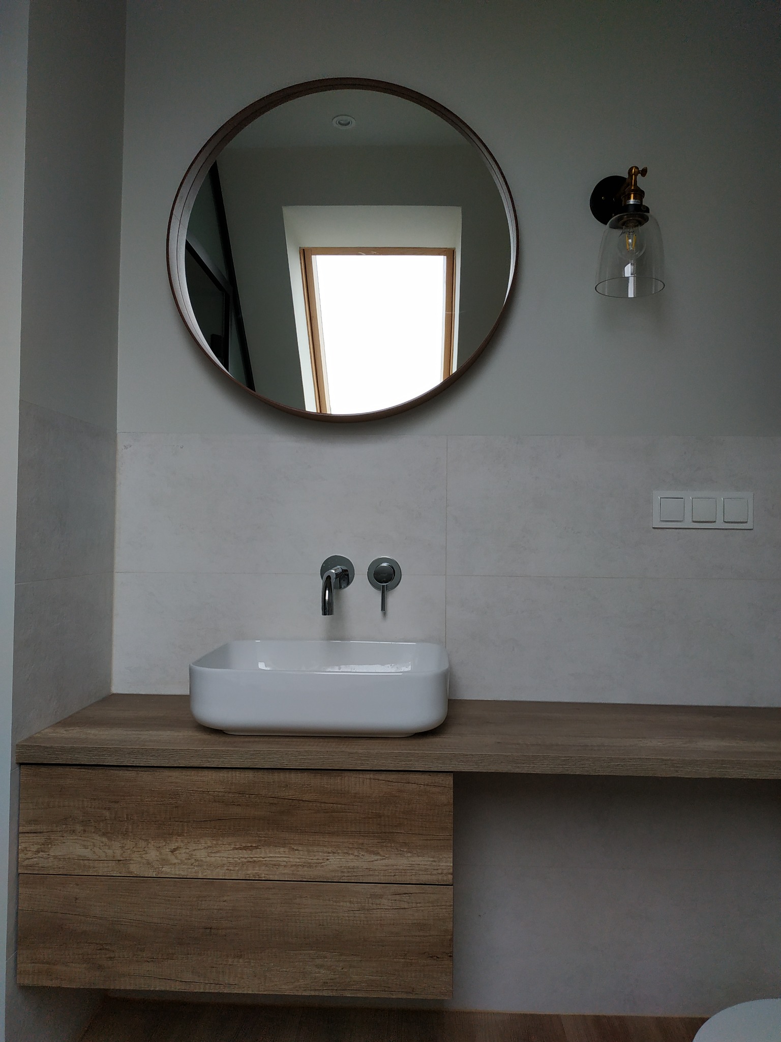 Nowoczesna łazienka z okrągłym lustrem, białą umywalką nablatową, drewnianą szafką pod umywalką i designerskim oświetleniem ściennym.