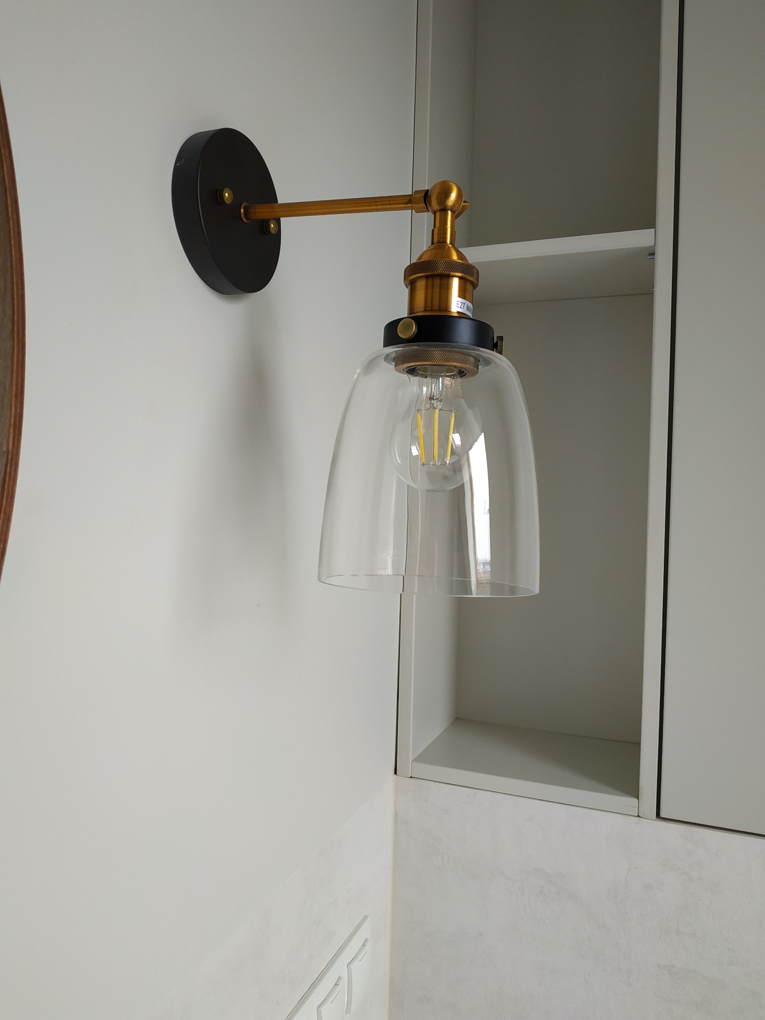 Ścienny kinkiet z czarną podstawą, złotym ramieniem i szklanym kloszem w łazience z szafkami w tle.