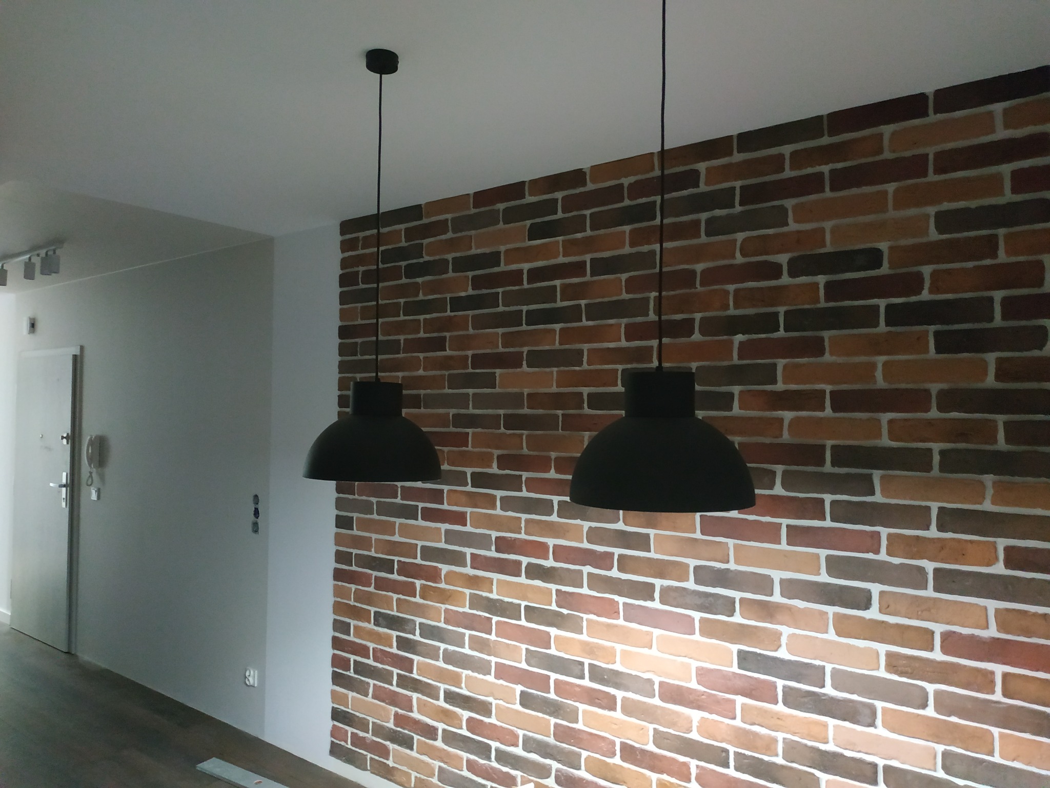 Ściana z dekoracyjnej cegły w odcieniach brązu i beżu, oświetlona dwoma czarnymi lampami wiszącymi w kształcie kloszy, widok na przedpokój z drzwiami wejściowymi.