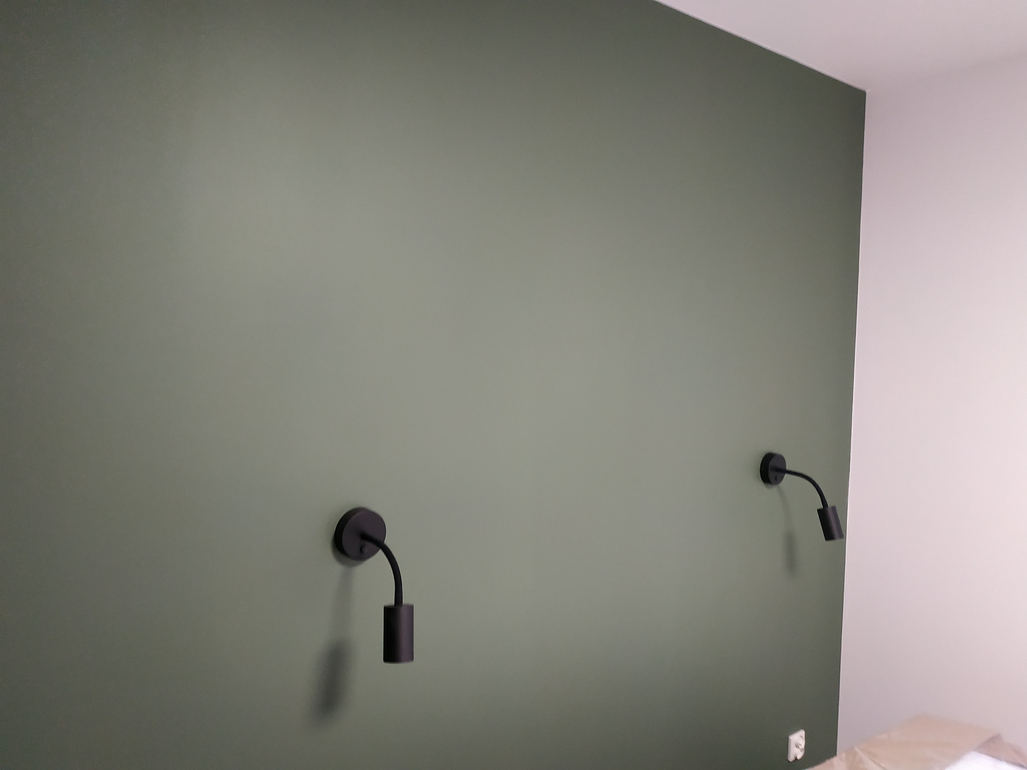 Ściana pomalowana na oliwkowy kolor z dwoma czarnymi kinkietami o minimalistycznym designie, widoczny fragment zabezpieczonej podłogi.