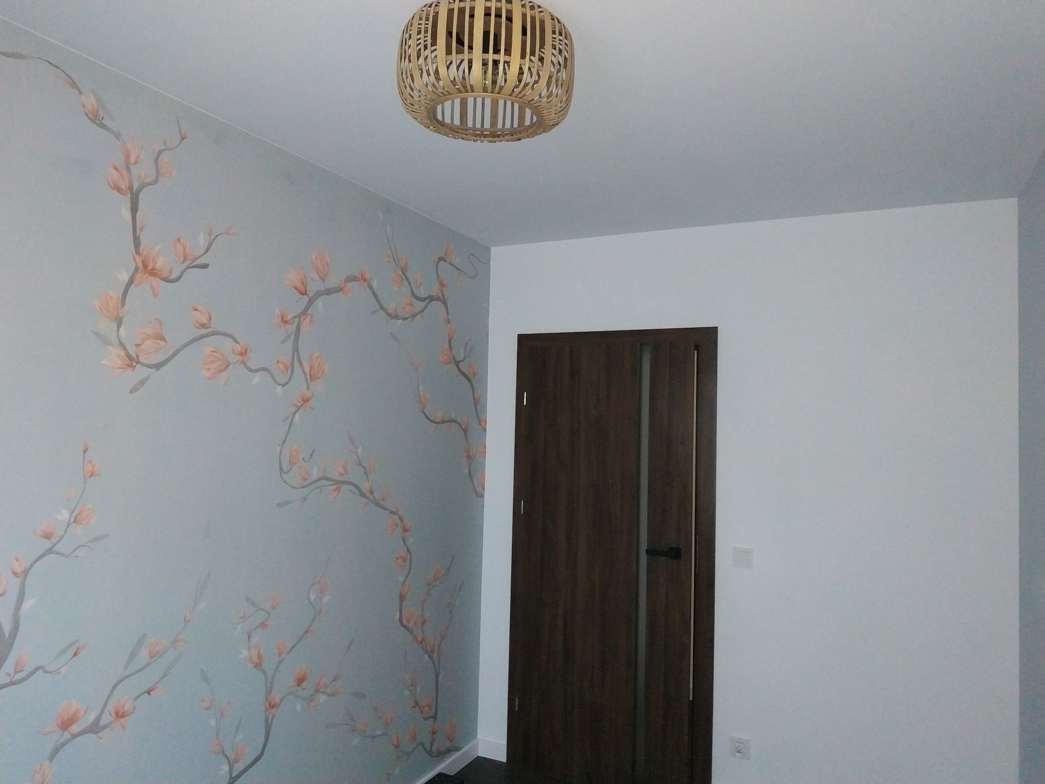 Ściana w pokoju z szarą tapetą w motywy kwitnących gałęzi, drewniana lampa wisząca i ciemne drzwi.